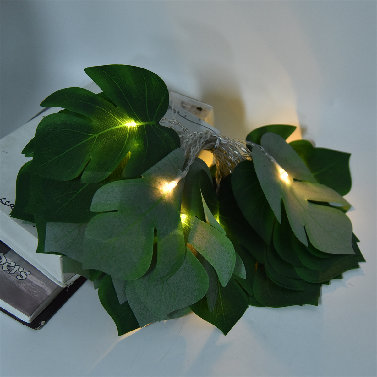 Eine Lichterkette mit grünen Kunstblättern und kleinen Lichtern, auf einer weißen Oberfläche.