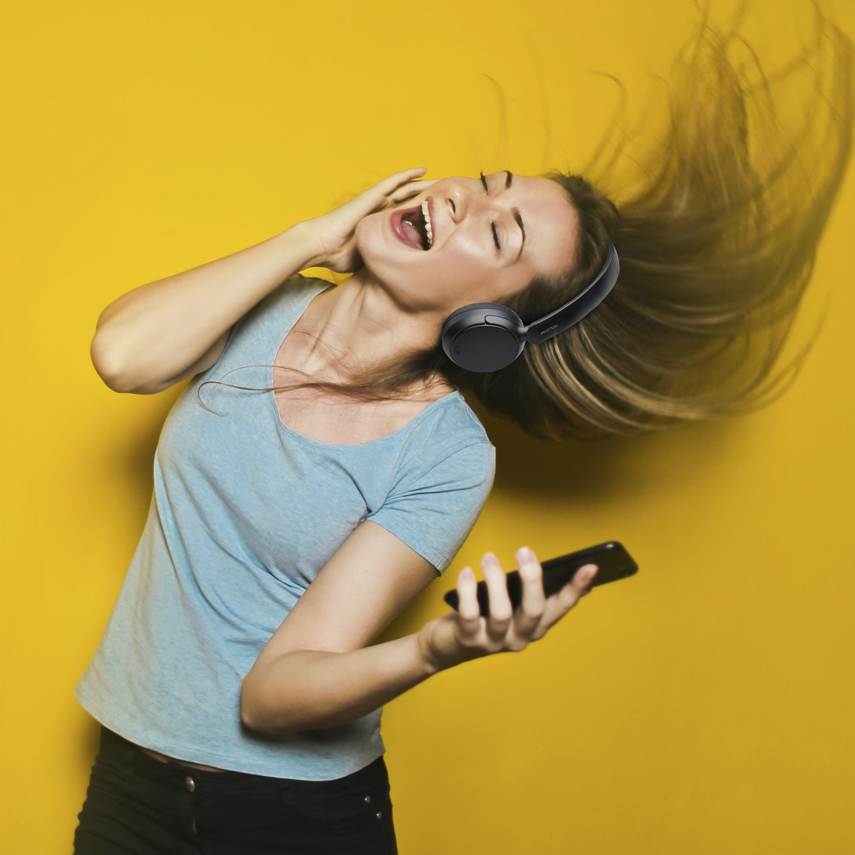 Mujer con auriculares cantando, sosteniendo teléfono. Fondo amarillo.