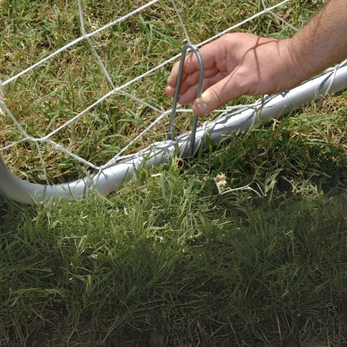Una mano che inserisce un paletto di metallo nel terreno vicino a una rete da calcio, erba verde.