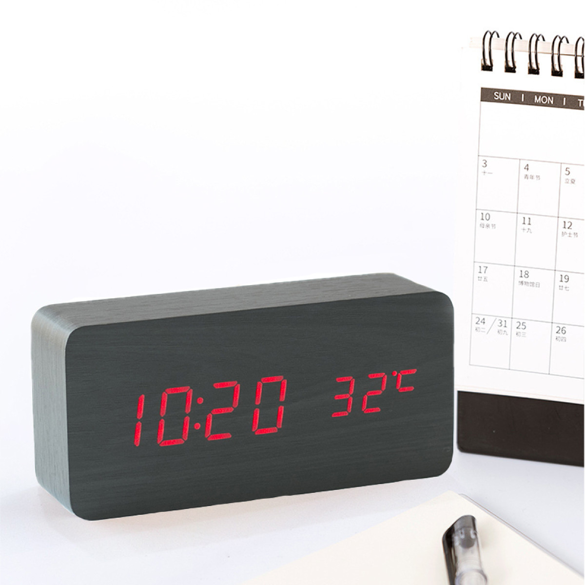 Czarny drewniany zegar cyfrowy z wyświetlaczem 10:20 i 32°C. Kalendarz widoczny w tle.
