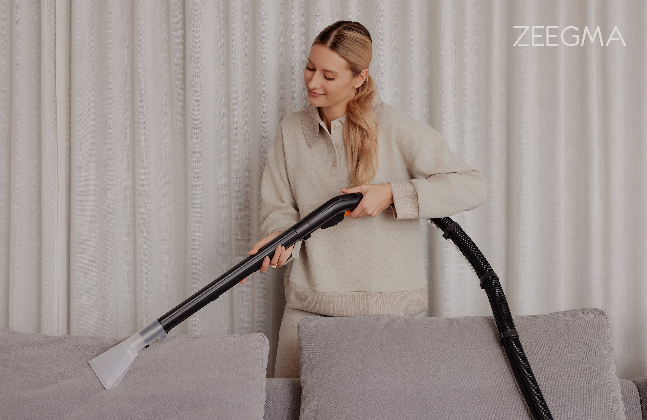 Frau benutzt einen Staubsauger auf einem grauen Sofa. Sie trägt beige Kleidung. Vorhänge im Hintergrund.