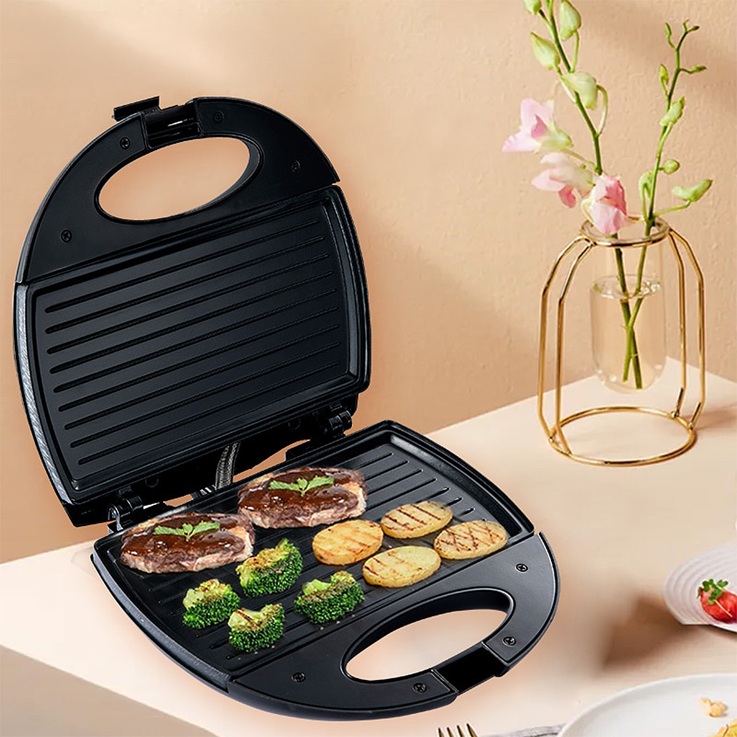 Ein offener schwarzer Sandwichmaker mit Essen, Brokkoli und Kartoffeln. Daneben eine kleine Vase mit rosa Blumen.