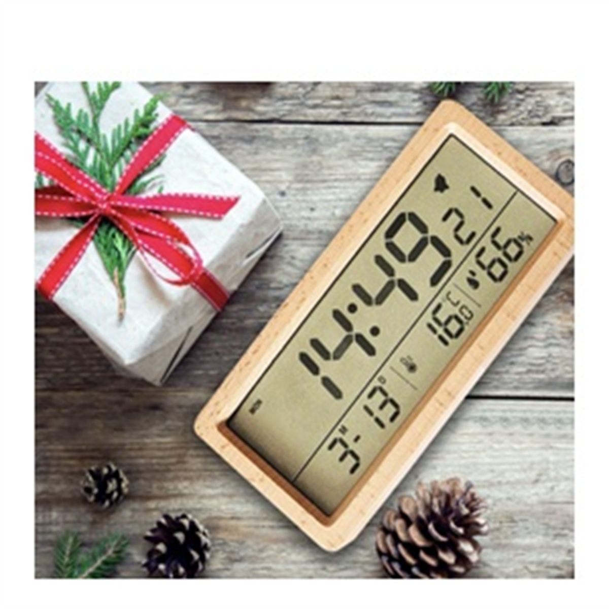 Digitaluhr mit Holzrahmen auf einem Holztisch. Eine Geschenkbox und Tannenzapfen sind in der Nähe.