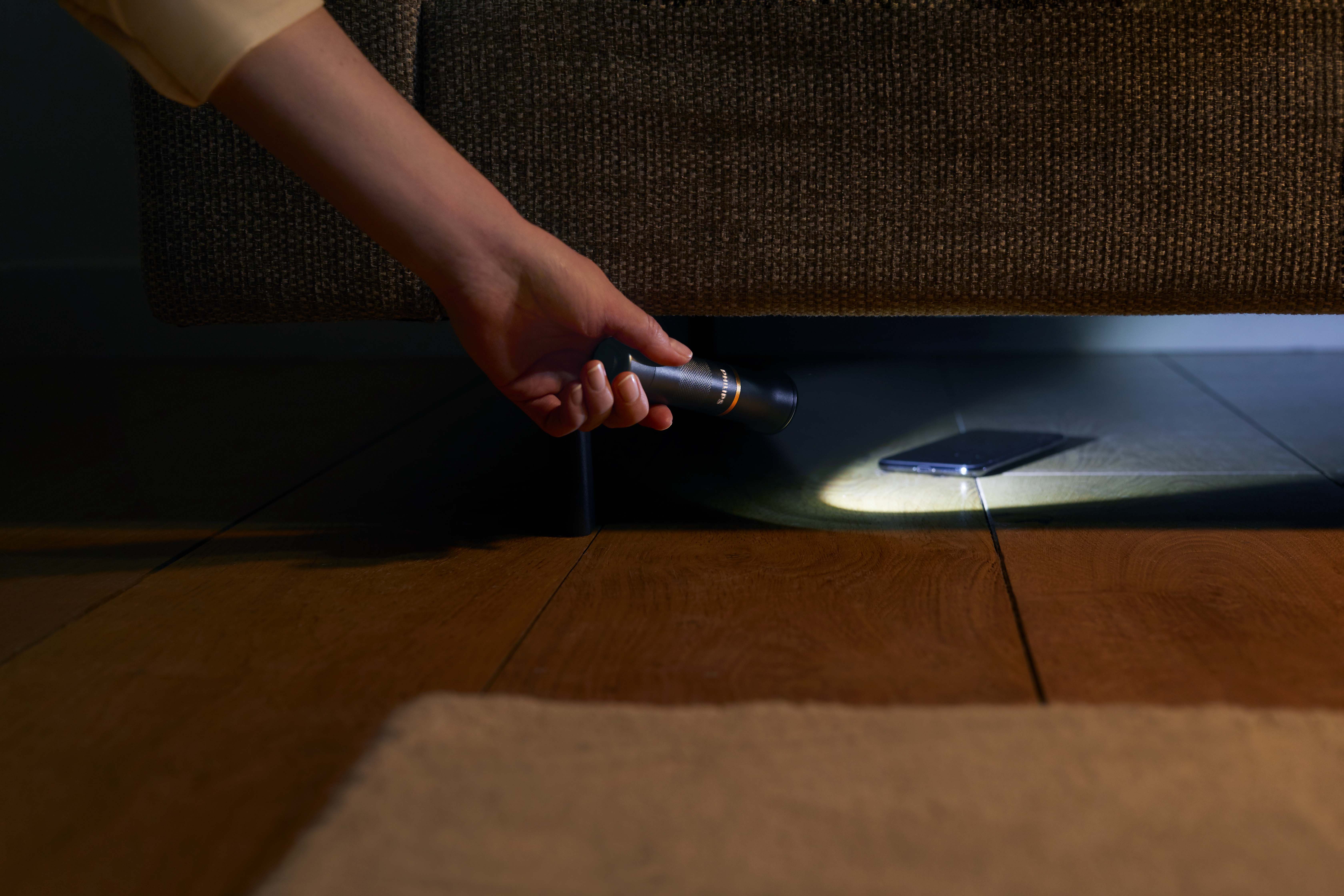 Hand hält eine Taschenlampe unter einem Sofa, sucht ein Telefon auf dem Boden.