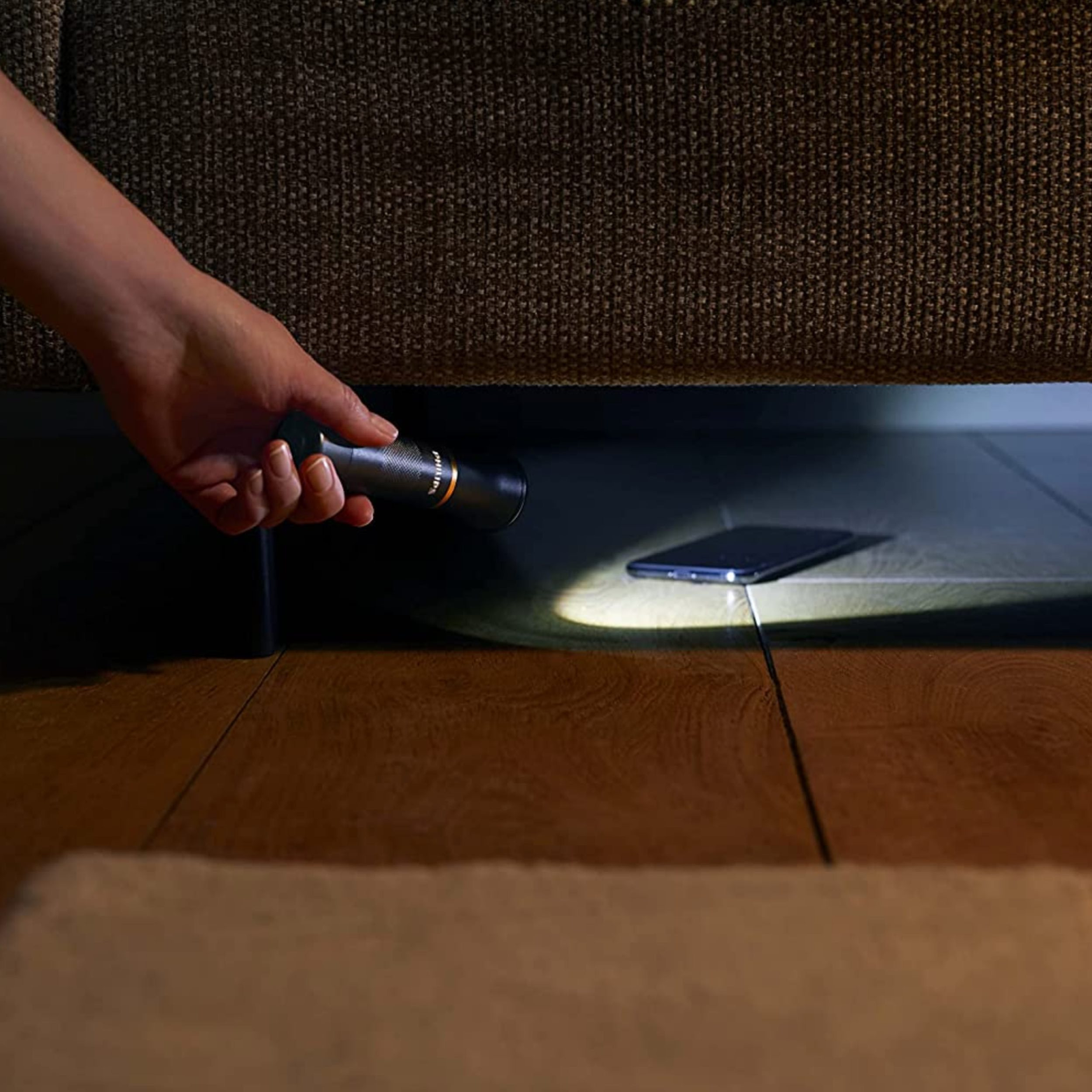 Hand mit Taschenlampe, die ein Telefon unter einem Sofa auf einem Holzboden beleuchtet.