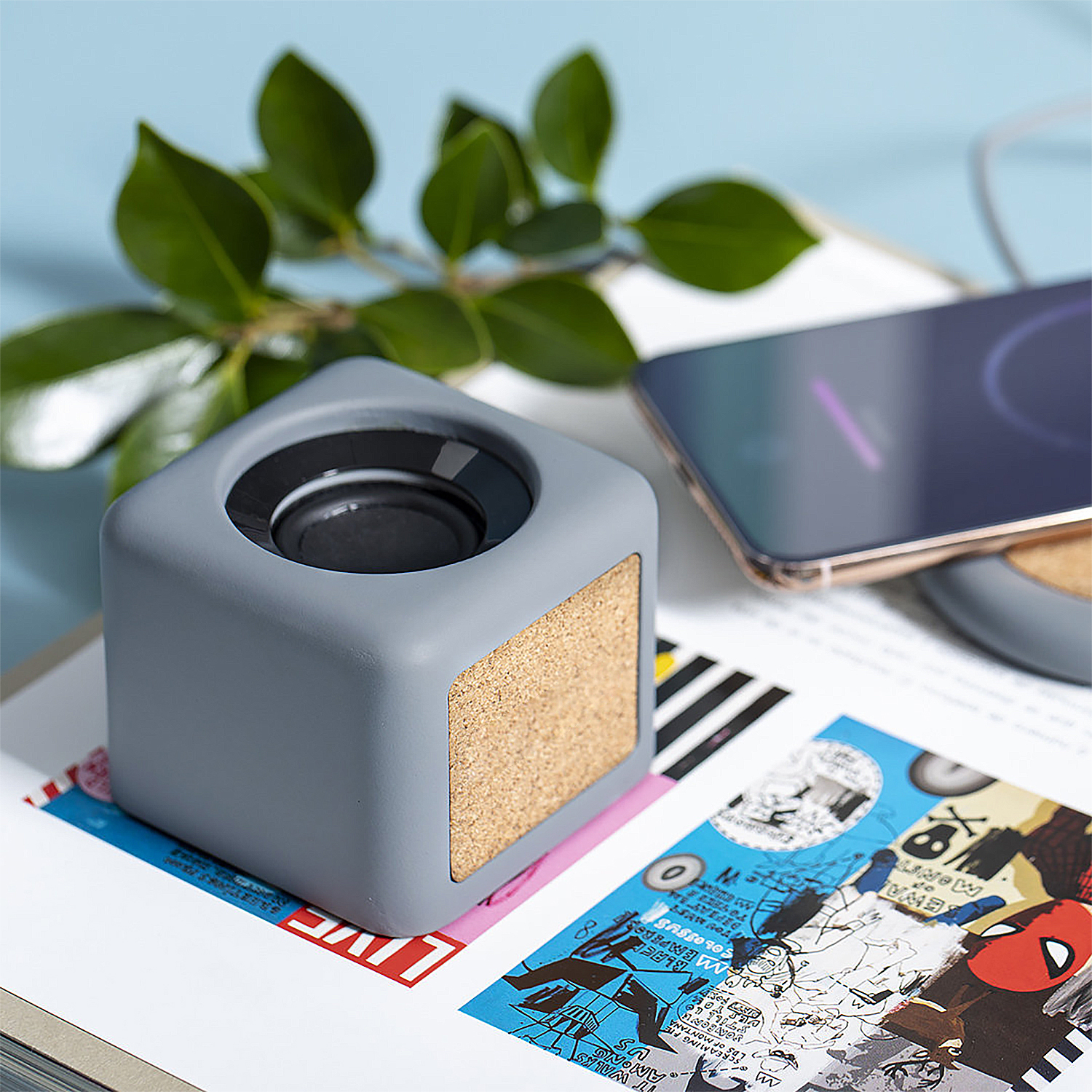 Un altoparlante grigio con accento in sughero è posizionato su una rivista colorata. Un telefono si carica sullo sfondo, con foglie verdi.