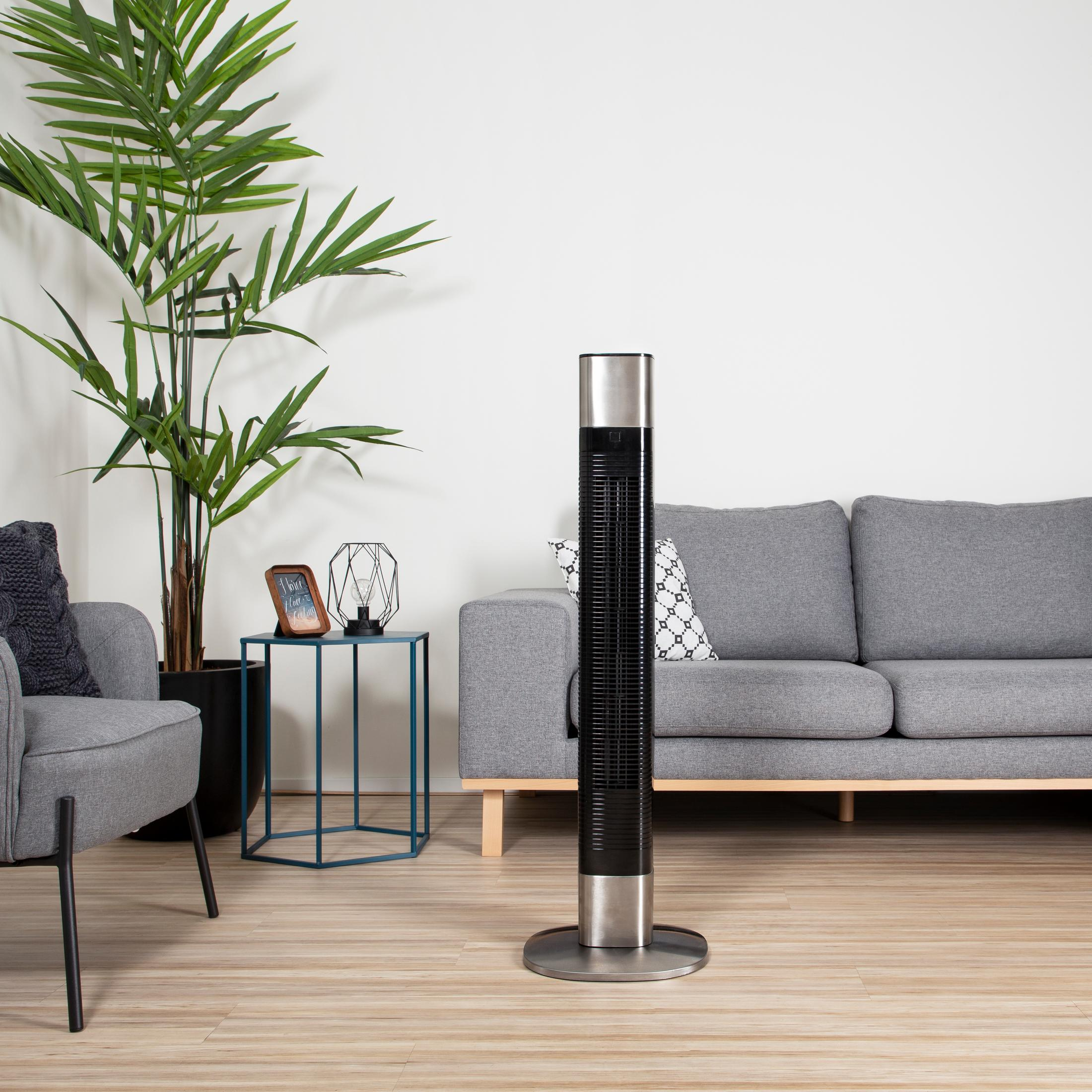 Wysoki czarny wentylator w pokoju. Szara sofa, fotel, stolik z dekoracjami i rośliną w tle.