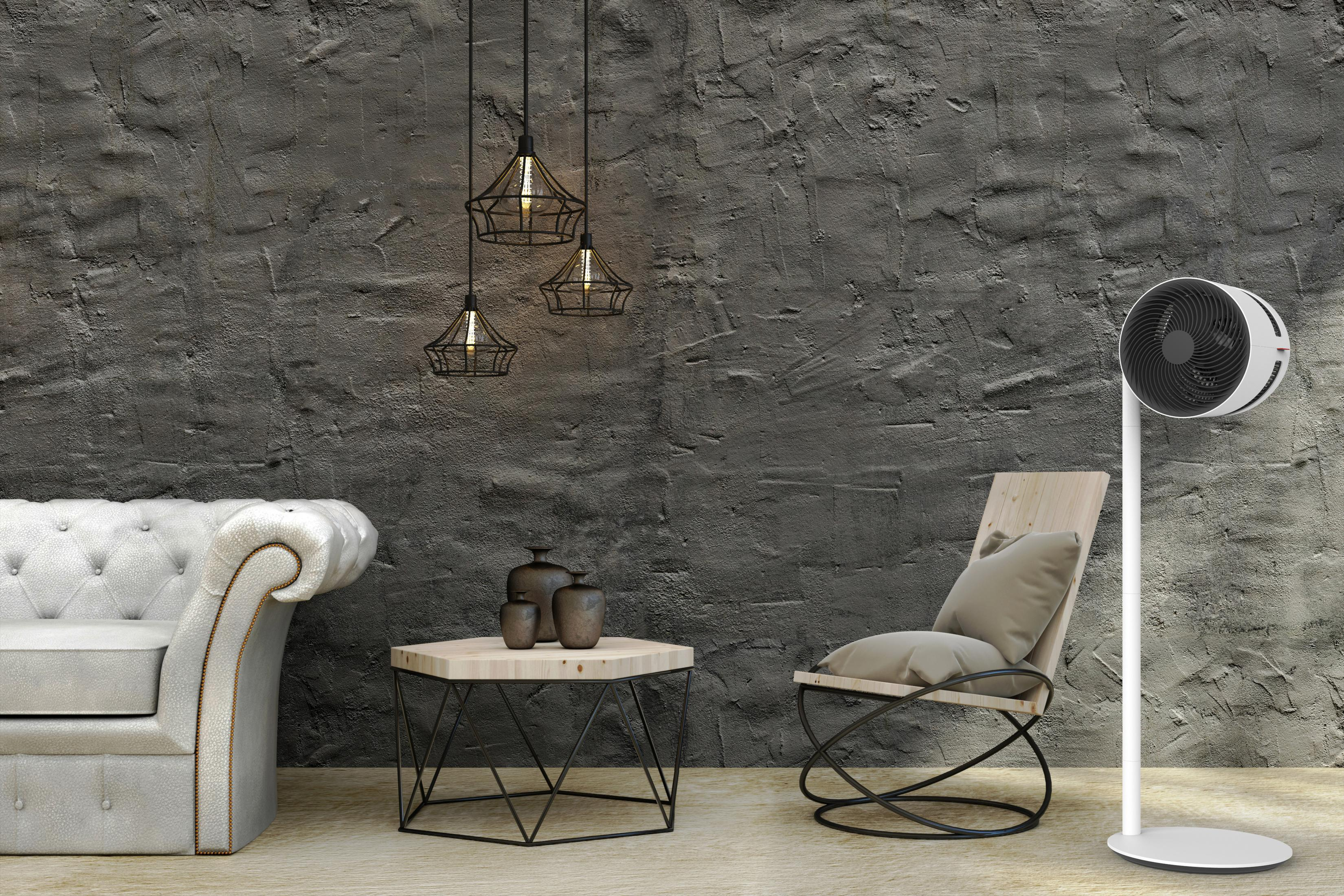 Scène de salon : canapé gris, table d'appoint avec décor, lampes noires, ventilateur sur pied blanc et chaise.