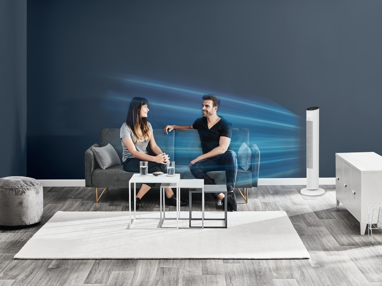 Dwie osoby na szarej kanapie. Wentylator wieje w ich stronę. Stoły i dywan przed nimi.