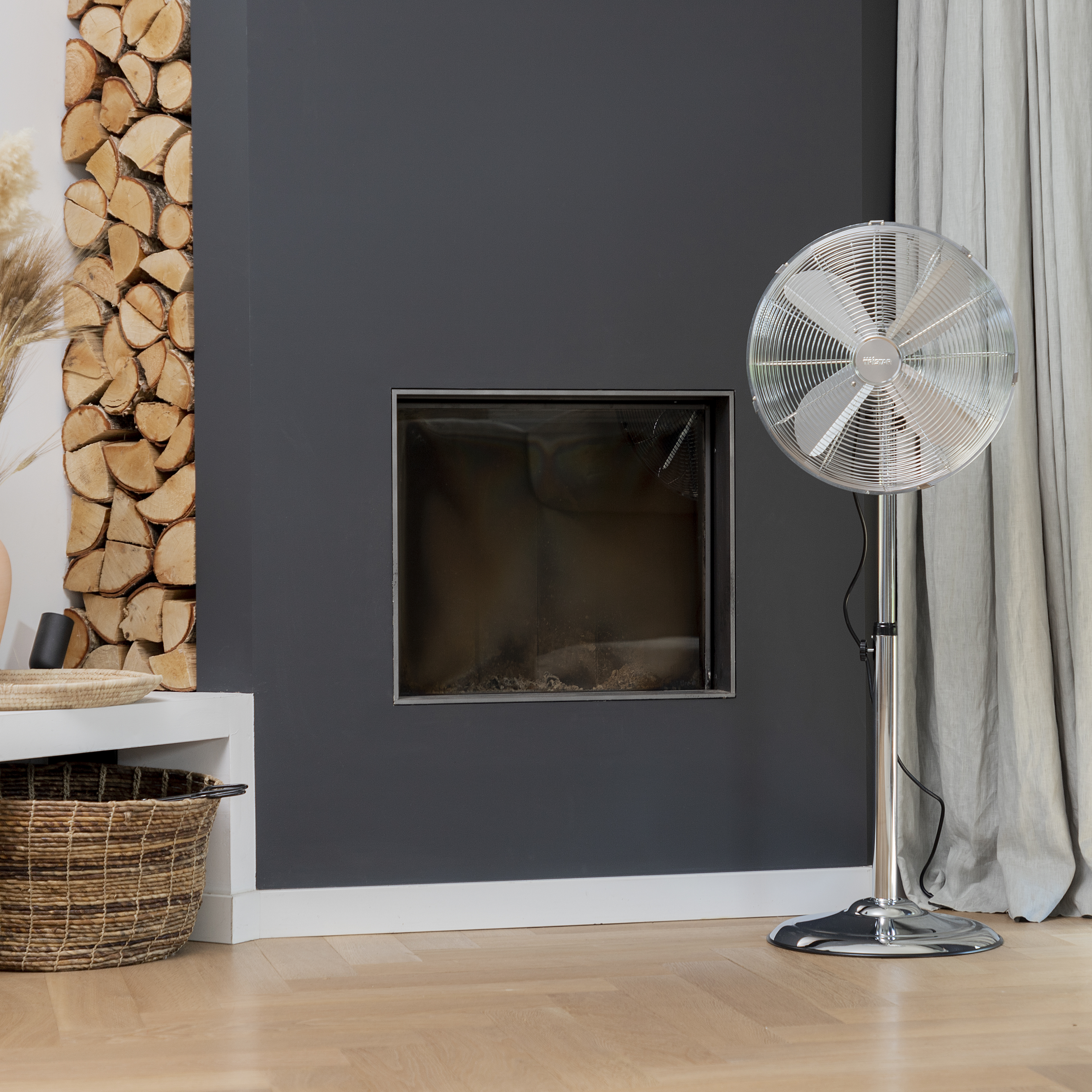 Ventilatore argentato su supporto, parete grigio scuro con camino, catasta di legna e tenda grigio chiaro.