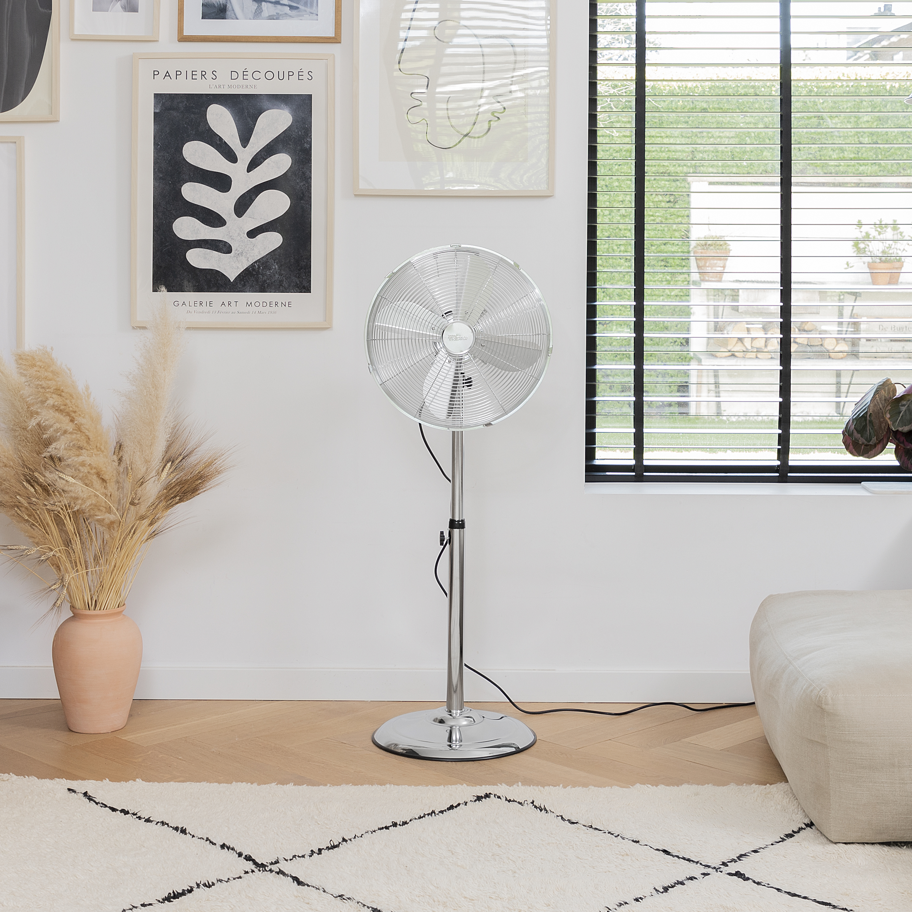 Ventilatore a colonna argentato in un soggiorno moderno. Arte sulla parete, tappeto e divano beige visibili.