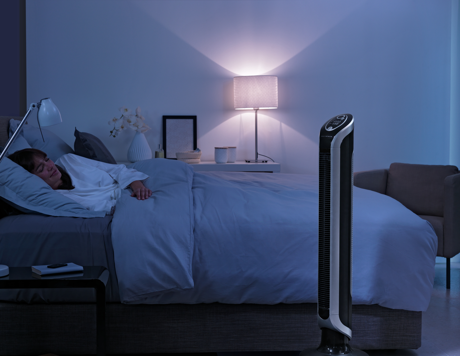 Scena w sypialni w nocy z kobietą śpiącą w łóżku i wysokim wentylatorem działającym w rogu.