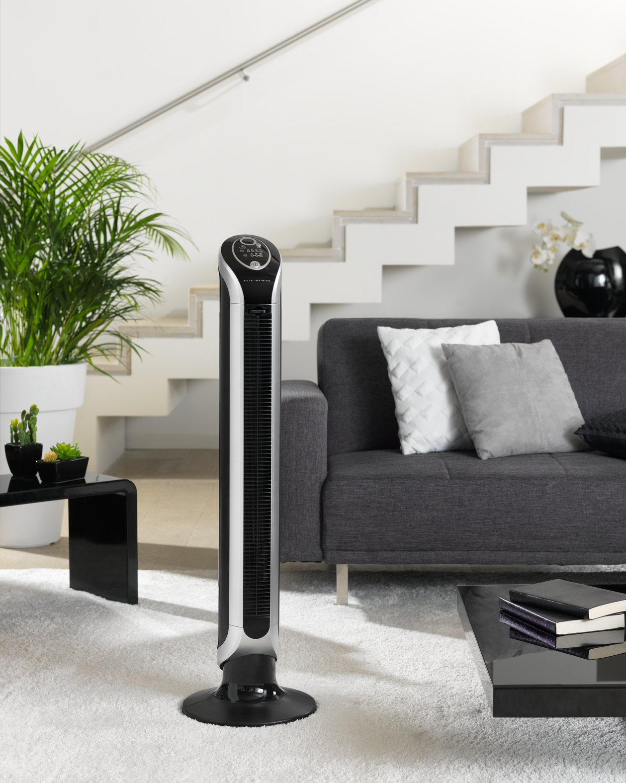 Czarny i srebrny wentylator wieżowy stoi na białym dywanie w salonie, w pobliżu szarej sofy i schodów.