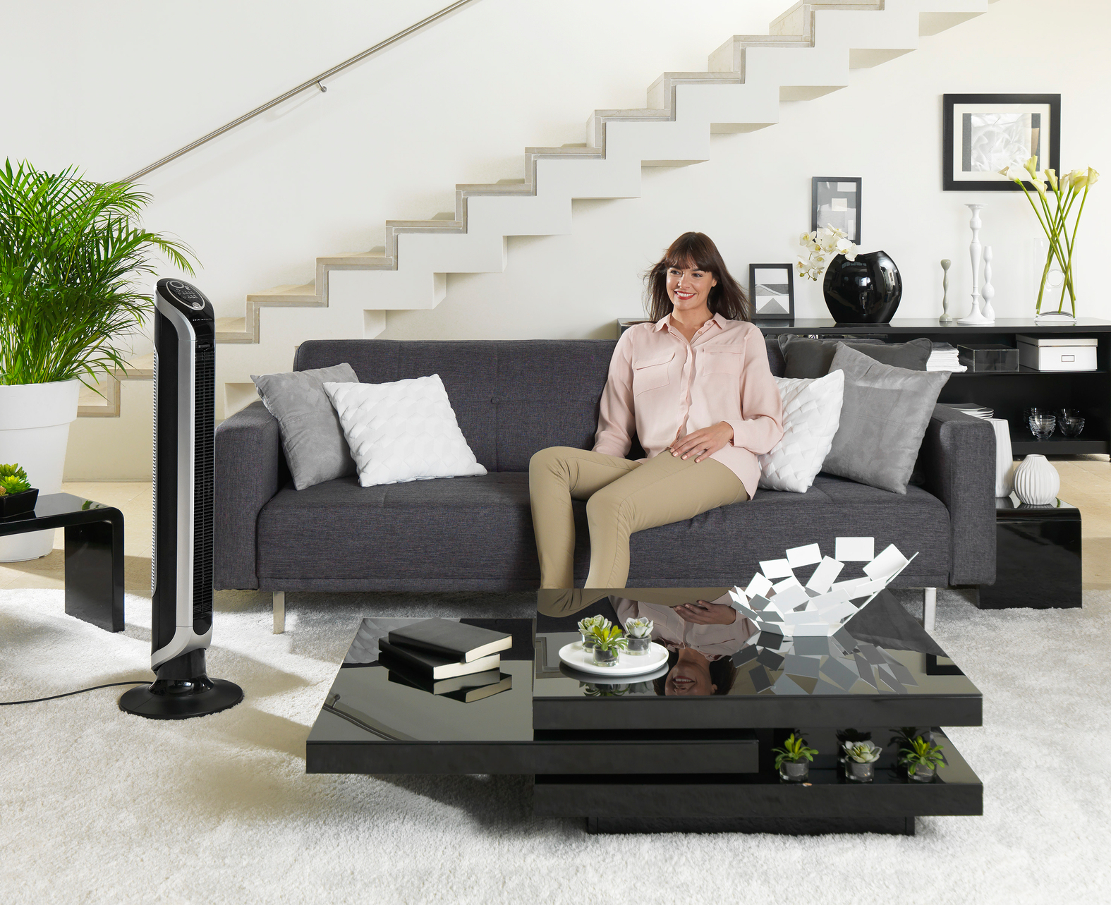 Kobieta siedzi na szarej sofie. Czarny wentylator po lewej. Czarny stół z przodu.