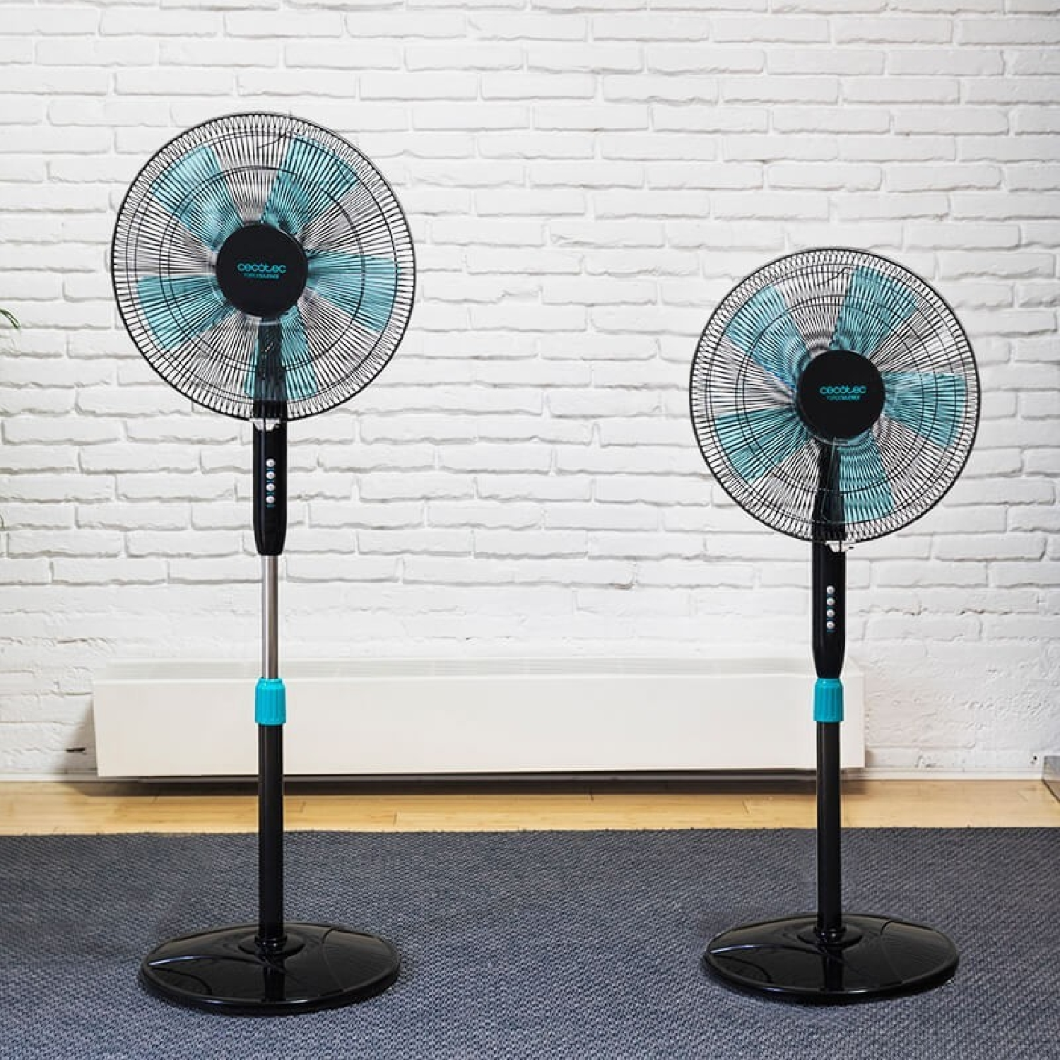Dos ventiladores de pie negros y turquesa, vista frontal, en una habitación con fondo de pared de ladrillo blanco.