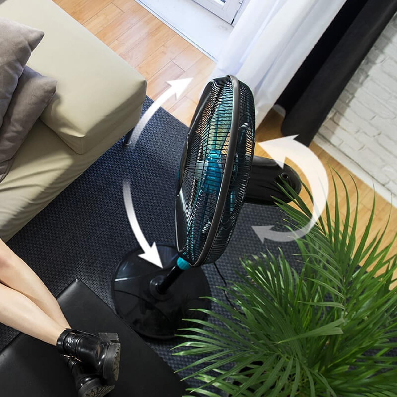 Un ventilador de pie negro y verde azulado con una base negra, junto a una planta verde y las piernas de una persona descansando.