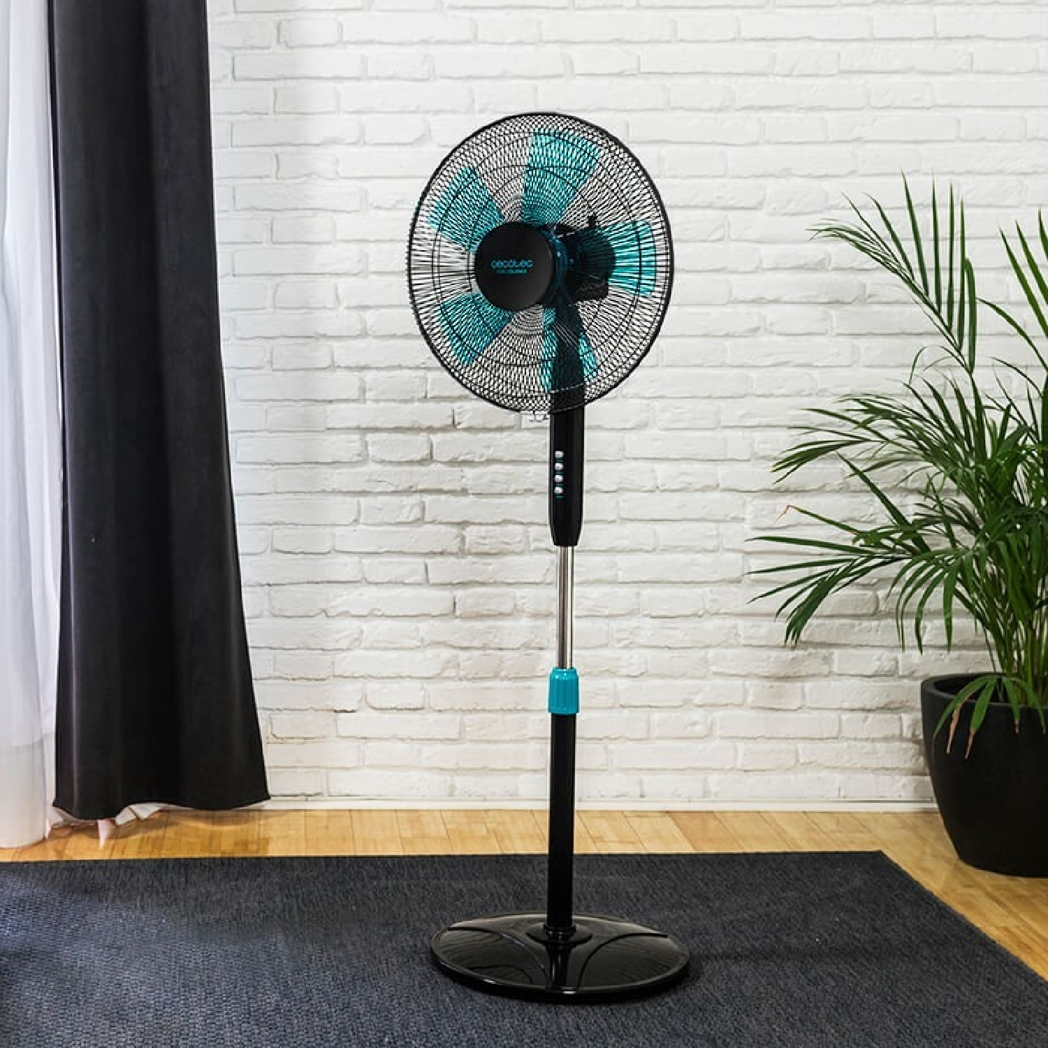 Un ventilador de pie negro y turquesa en una habitación con pared de ladrillo y planta.