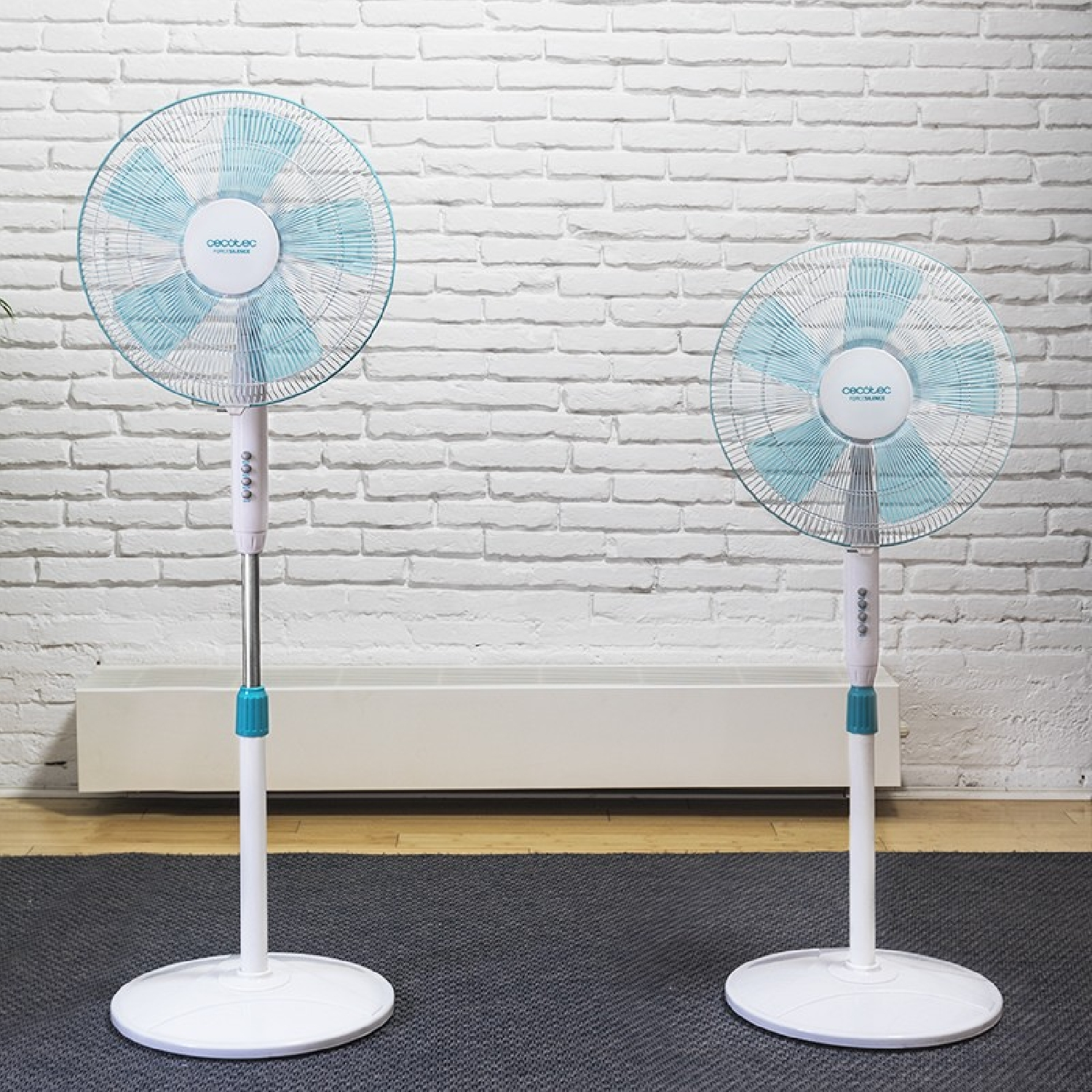 Dos ventiladores de pie blancos y azules. Los ventiladores están contra una pared de ladrillo, sobre una alfombra gris.