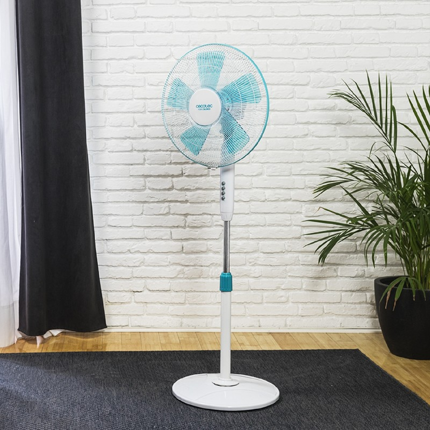 Un ventilador de pie con aspas azules, junto a una planta y una pared de ladrillo.
