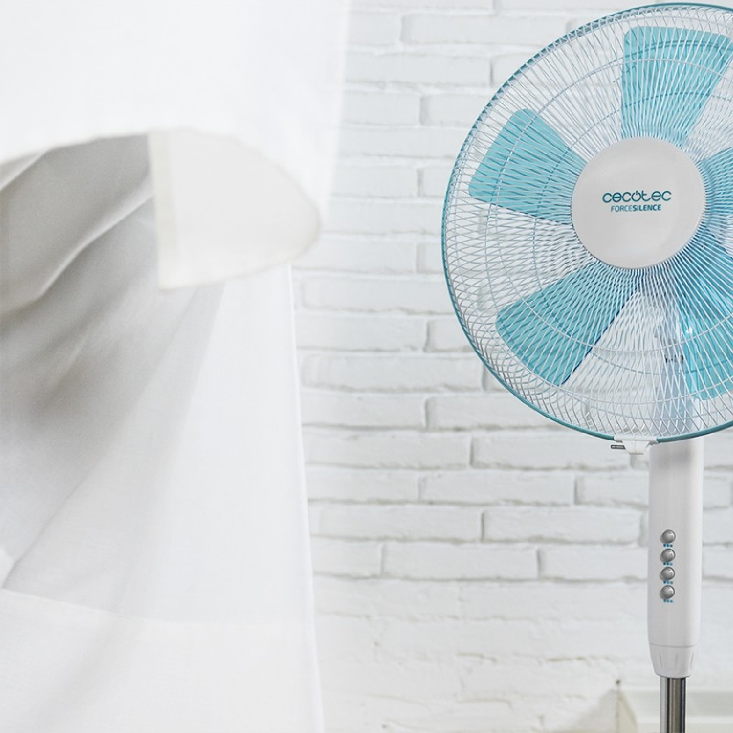 Un ventilador de pie azul y blanco está junto a una cortina blanca y una pared de ladrillo blanco.