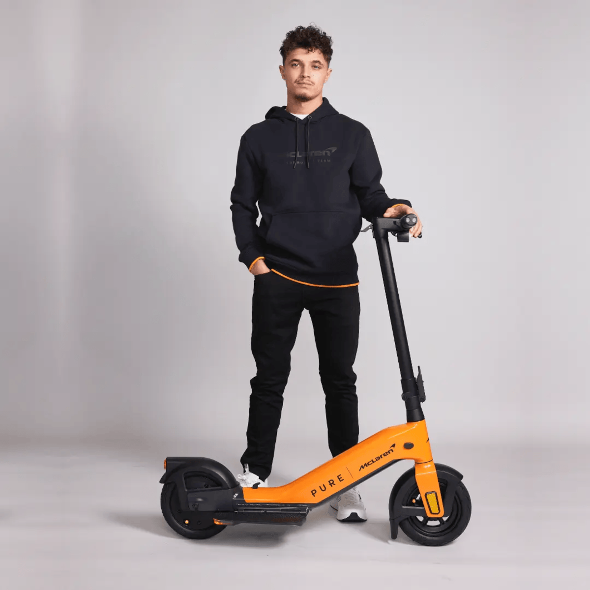 Ein Mann steht neben einem orangenen Roller, er trägt Schwarz. Auf dem Roller steht Pure McLaren.
