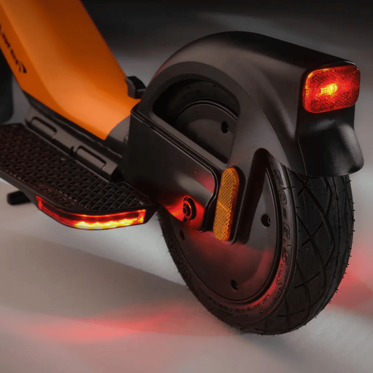 Rückseite des Elektrorollers, schwarz und orange. Rote Lichter hinten und an den Seiten. Schwarzer Reifen auf hellem Hintergrund.