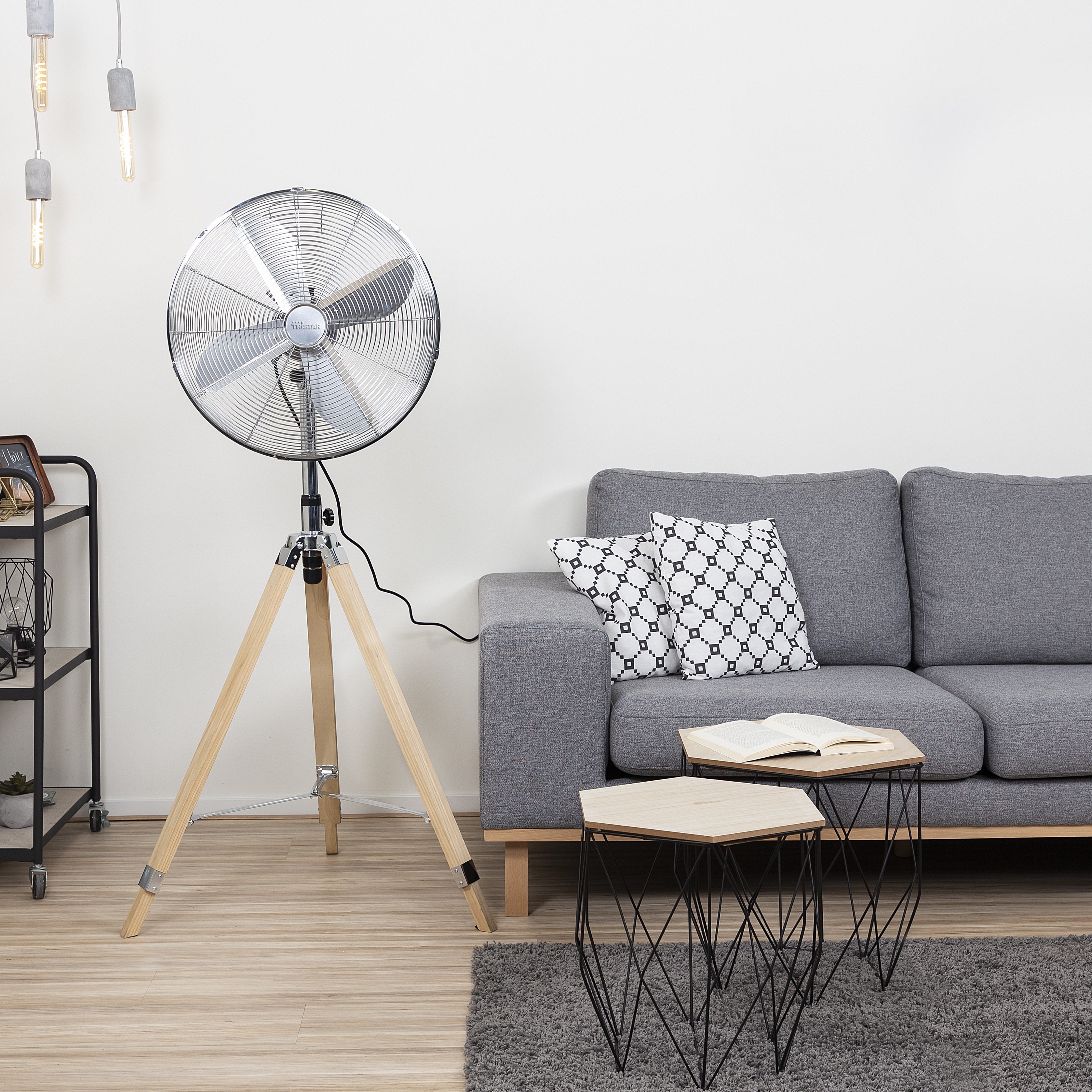 Un ventilatore argentato su gambe di legno vicino a un divano grigio e tavolini.