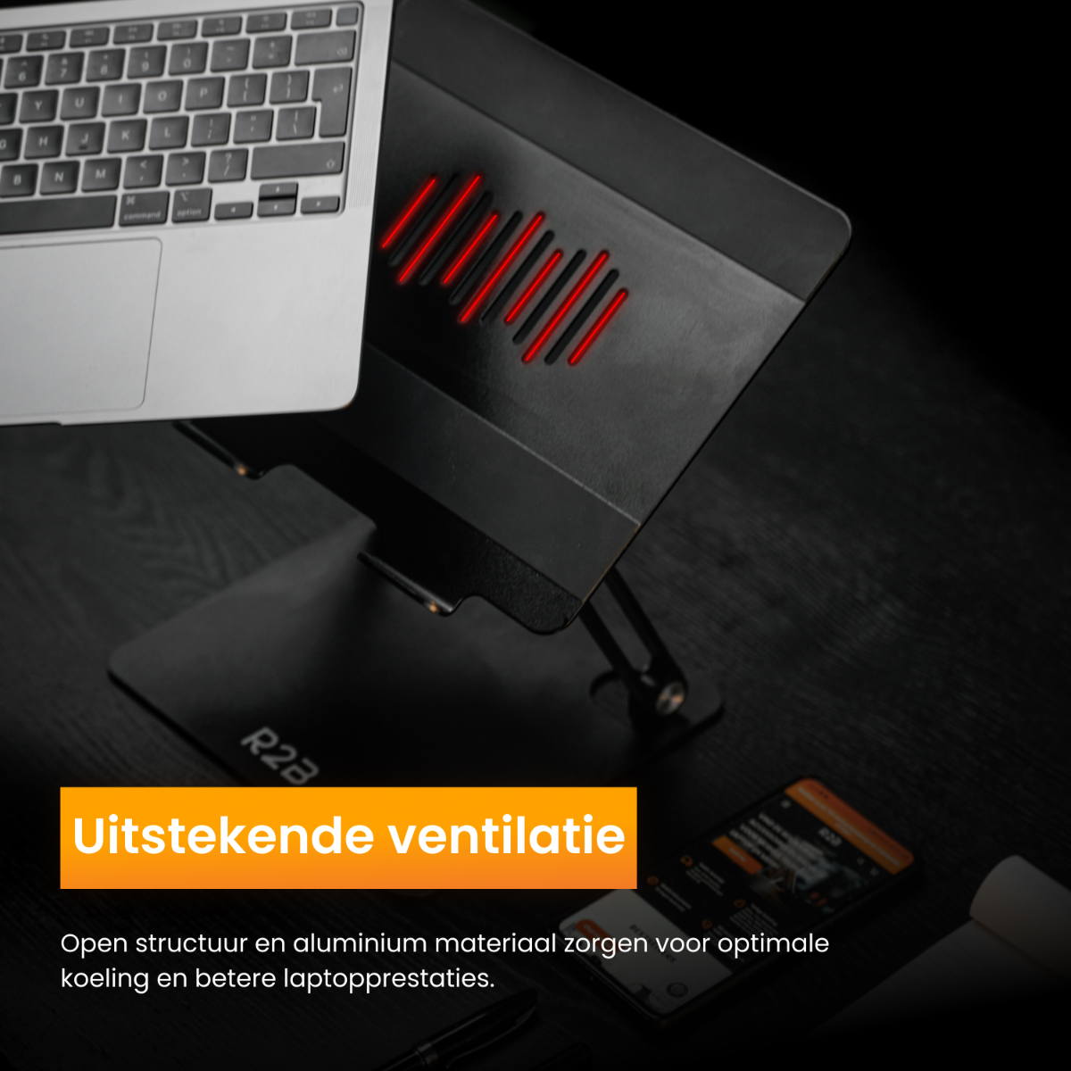 Laptop op zwarte standaard met rode ventilatie, telefoon op tafel, alles op een donker oppervlak.