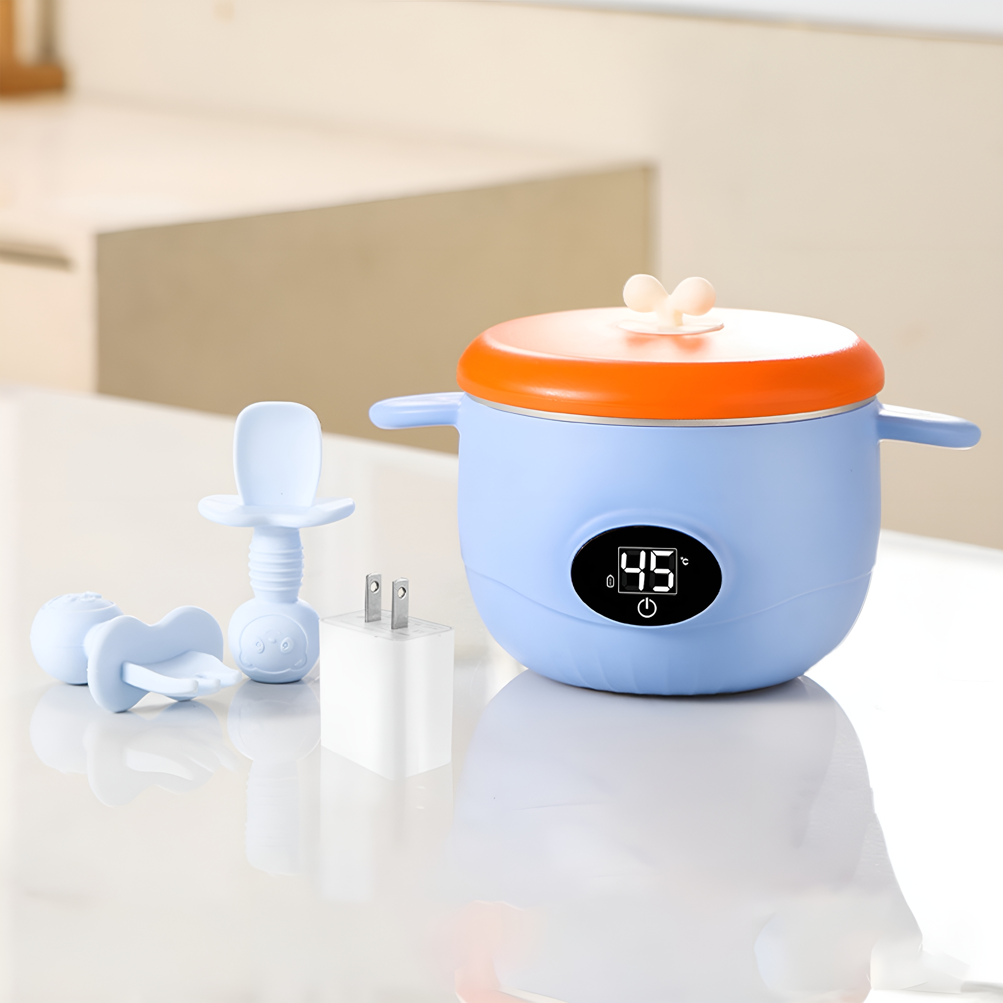Ein blau-orangener Babykostwärmer mit Löffeln und Stecker, alles auf weißer Oberfläche. Display zeigt 45.