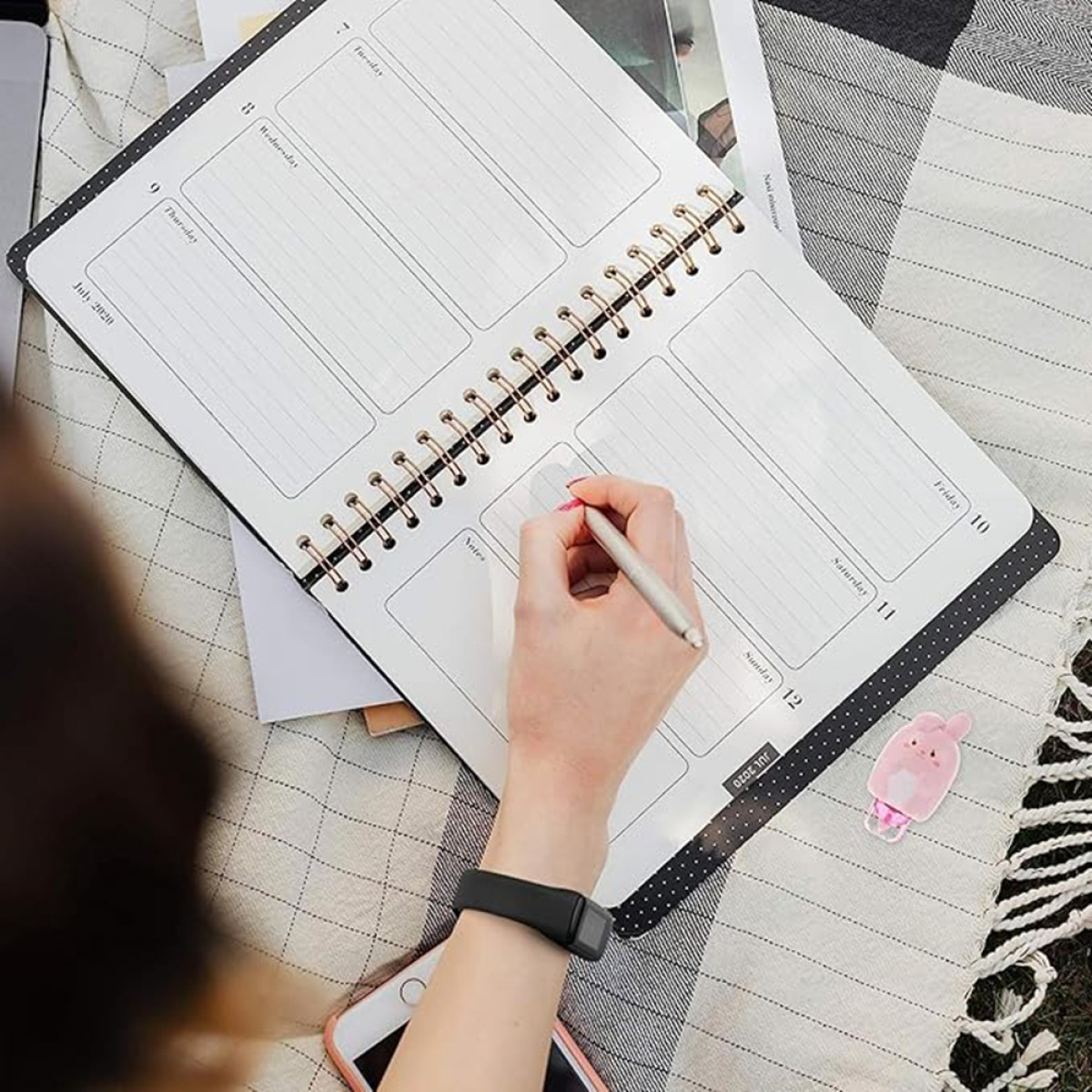Kobieta pisząca w planerze długopisem. Widoczny zegarek, telefon i naklejka.