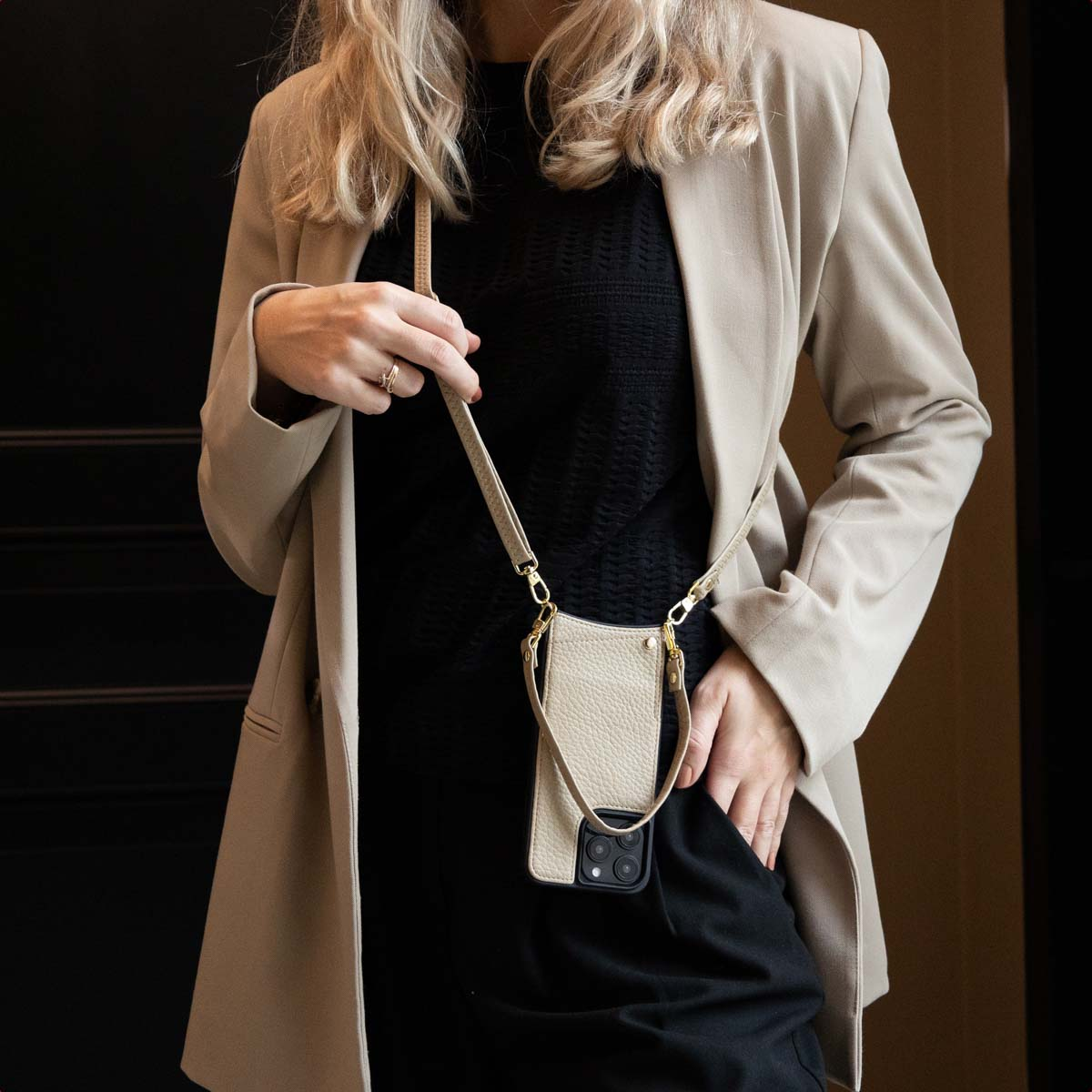 Una donna in blazer tiene un telefono in una custodia beige con un cinturino. È vicino a una porta nera.