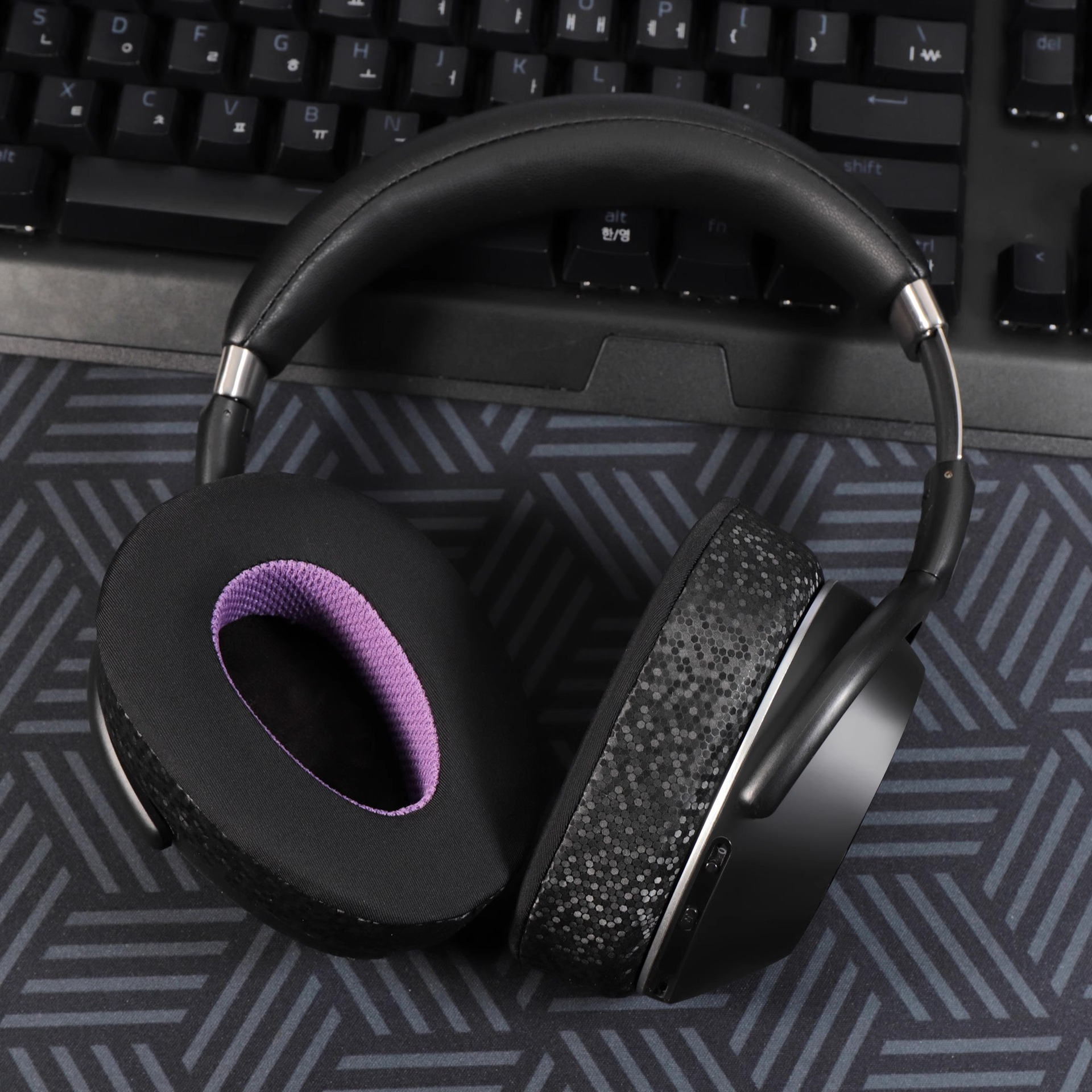 Czarne słuchawki z fioletowymi nausznikami, na teksturowanej powierzchni. Srebrne akcenty. Z tyłu czarna klawiatura.
