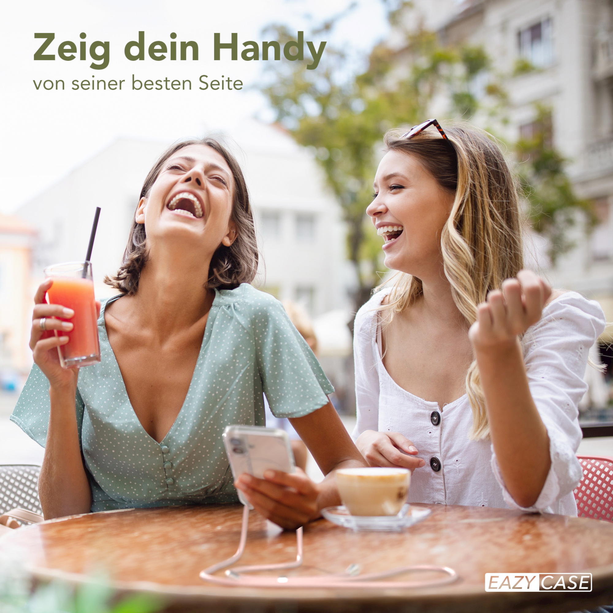 Zwei lachende Frauen, eine hält ein Telefon mit Band, sitzen an einem Tisch mit Getränken.