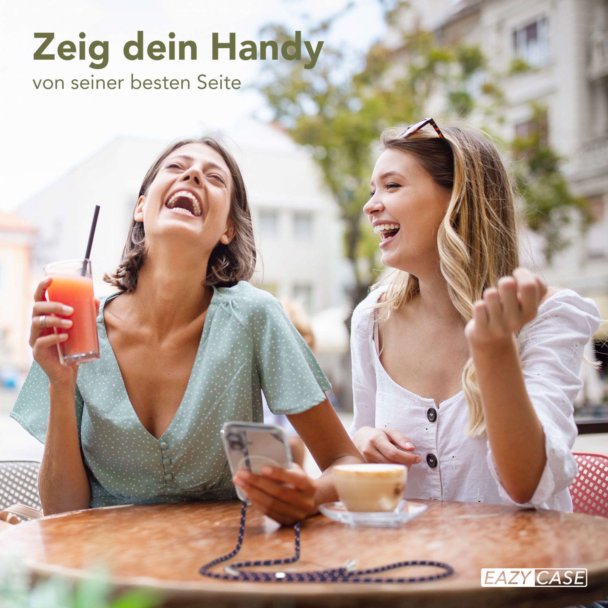 Zwei Frauen lachen, halten Getränke und ein Telefon mit blauem Schlüsselband. Café im Freien.