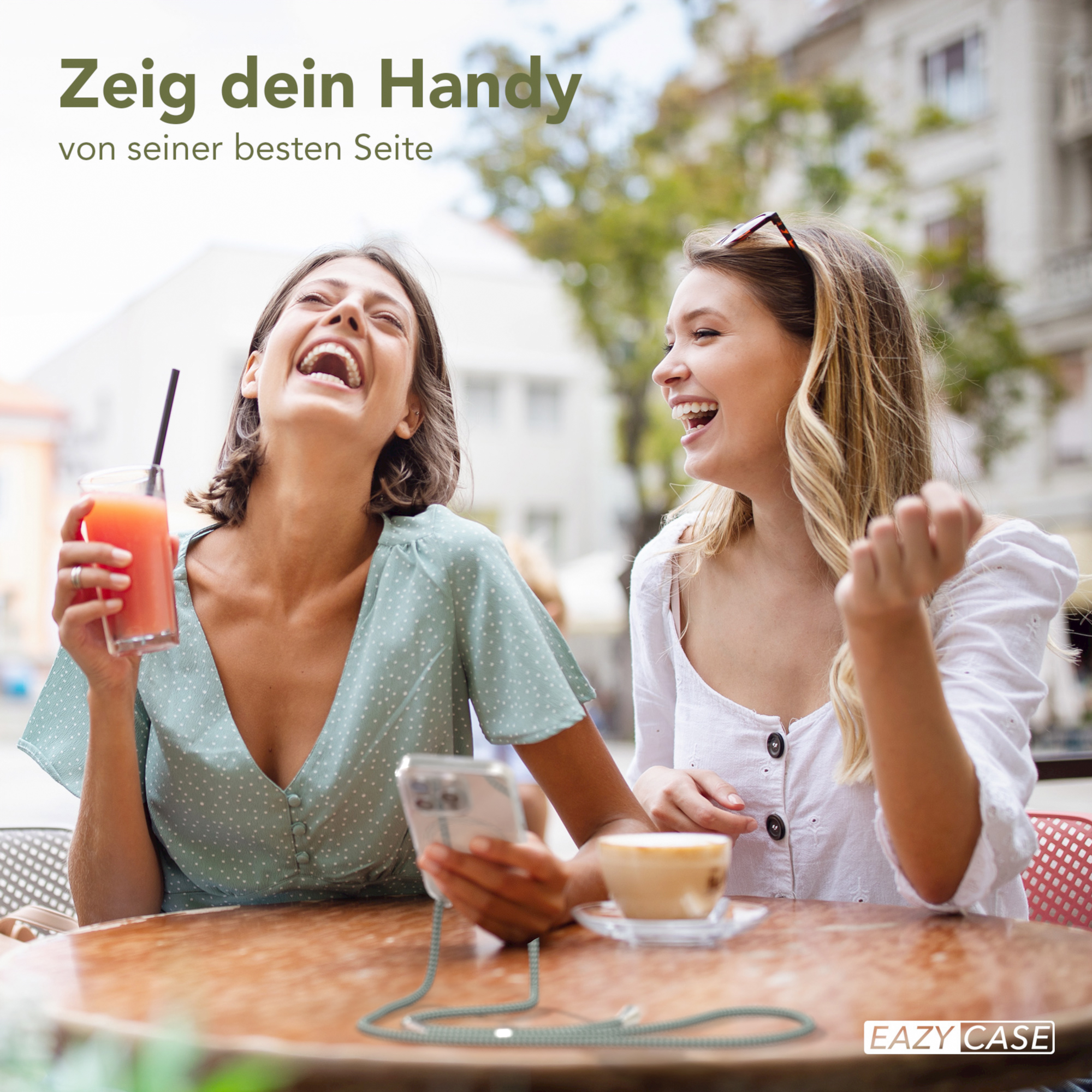 Zwei lachende Frauen im Freien, eine hält ein Telefon. Ein Saftglas und eine Kaffeetasse stehen auf dem Tisch.
