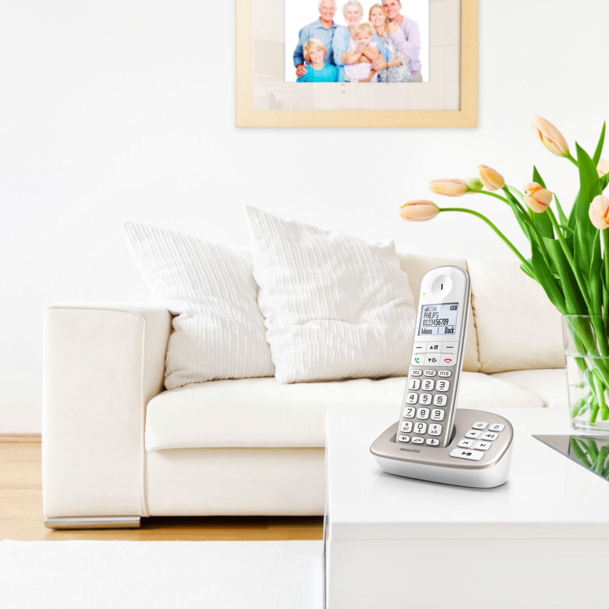 Weißes Zimmer mit weißem Sofa, Telefon auf einem Tisch und einem gerahmten Familienfoto. Blumen in einer Vase rechts.