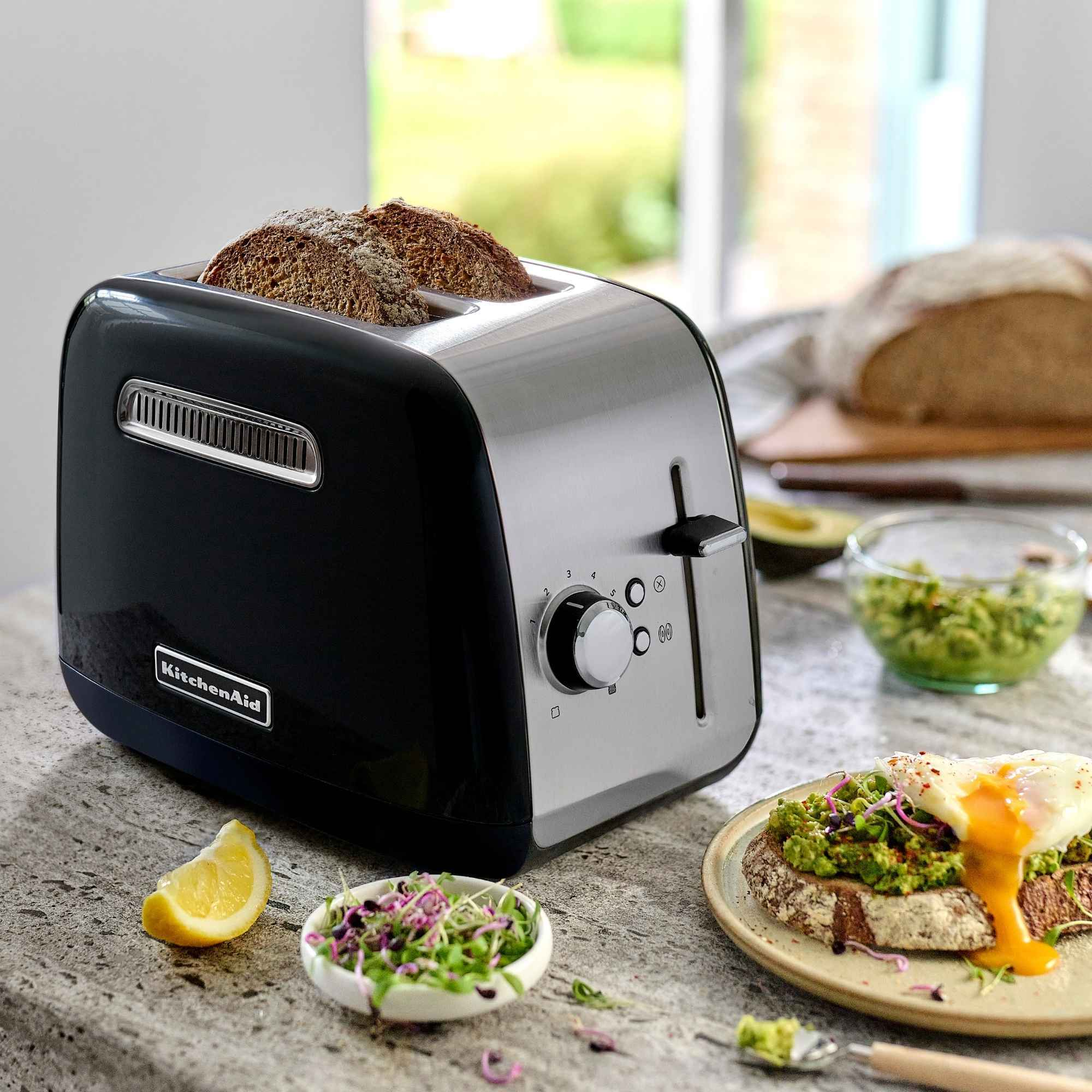 Black and silver toaster with two slices of toast inside. Avocado toast and ingredients on the side.