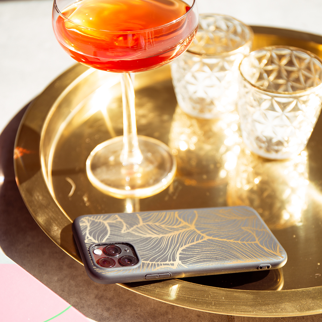 Ein Telefon mit schwarzer Hülle und goldenem Blattmuster auf einem goldenen Tablett mit Cocktail und Gläsern.