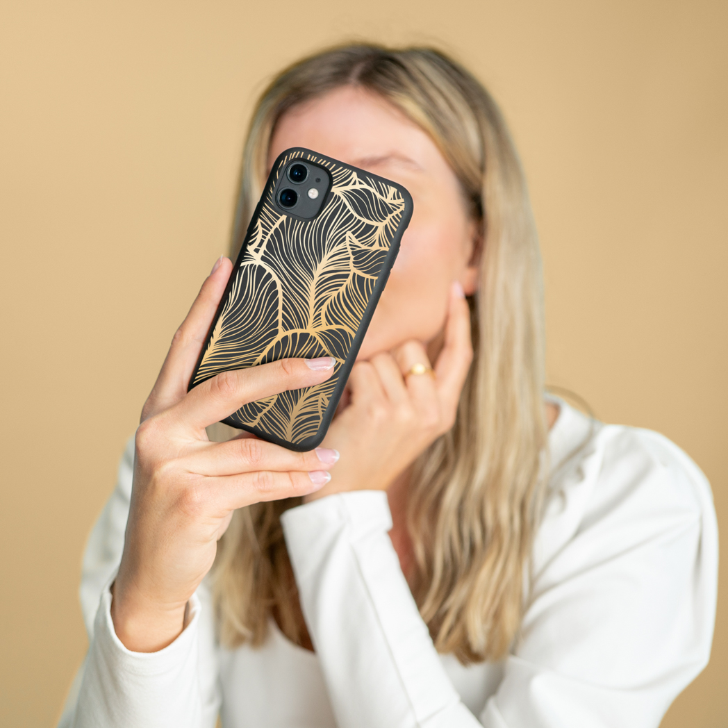 Frau hält schwarze Handyhülle mit goldenem Blattmuster, verdeckt ihr Gesicht.