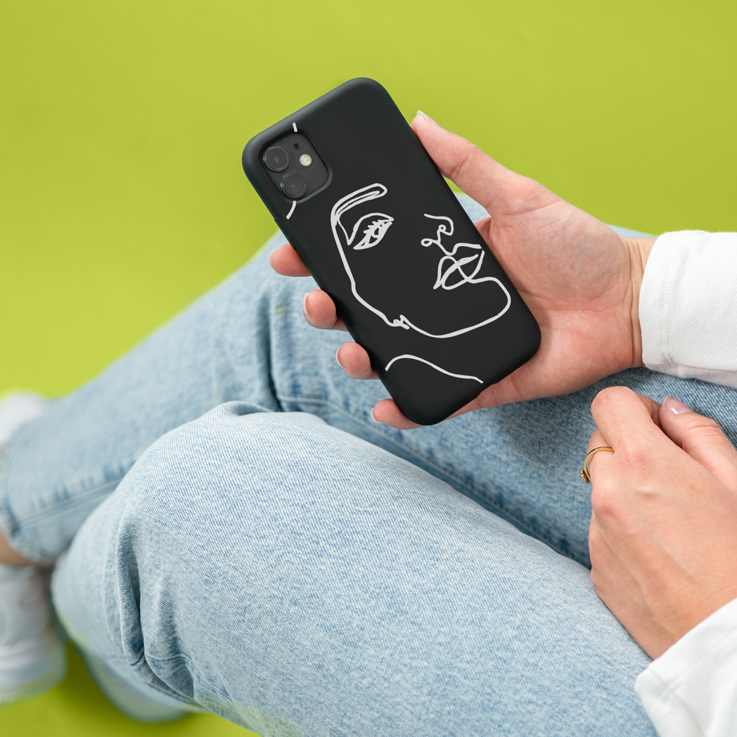 Vrouw in spijkerbroek houdt zwarte telefoonhoes vast met witte gezichtslijnkunst, groene achtergrond.