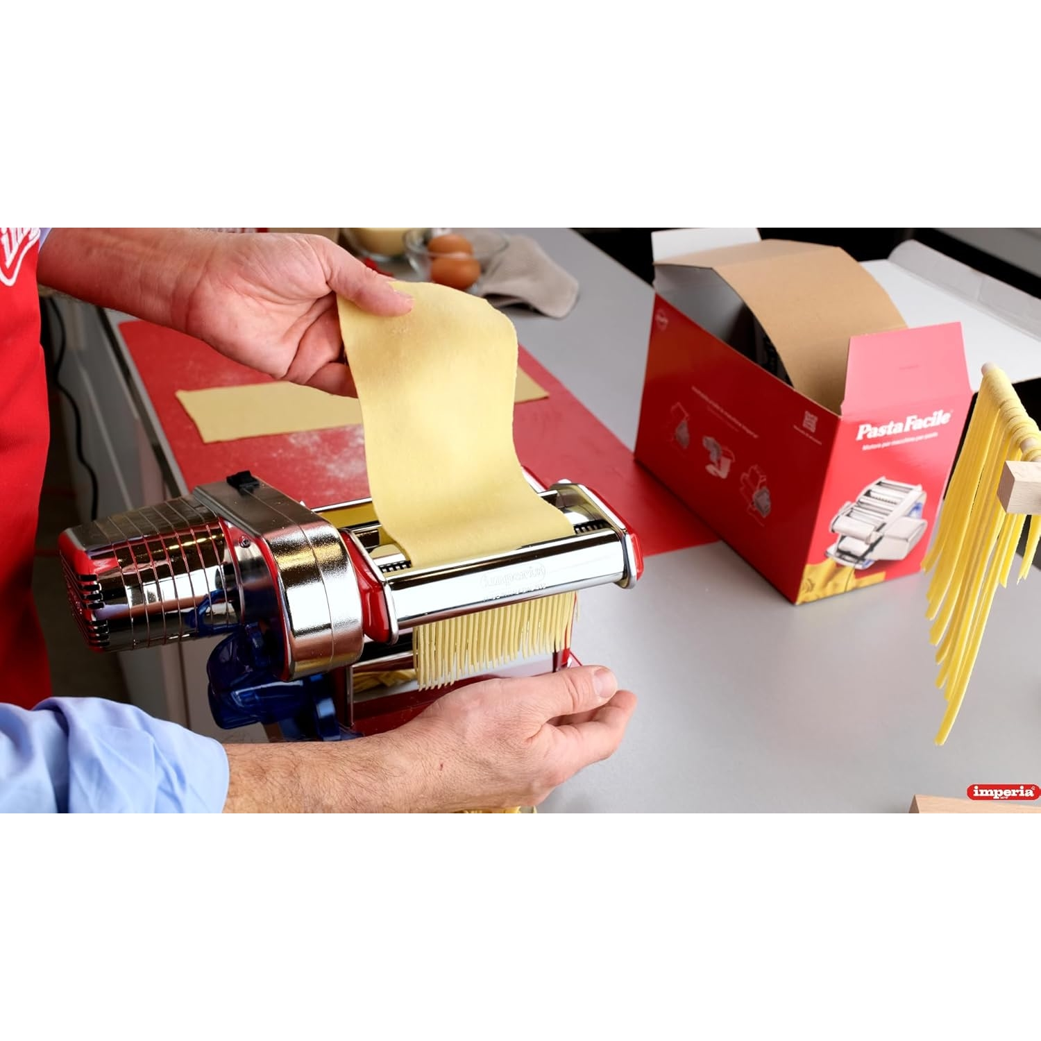 Mani che usano una macchina per la pasta per tagliare l'impasto. Una scatola rossa e un ripiano di pasta sullo sfondo.