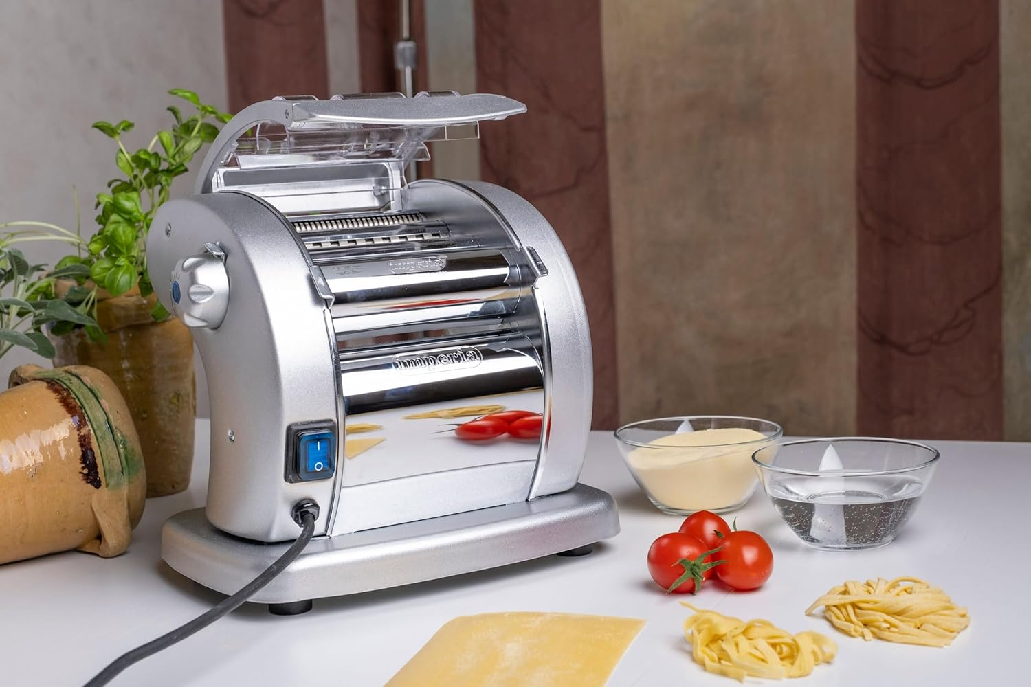 Macchina per pasta con ingredienti su superficie bianca, tra cui pomodori, basilico e pasta.