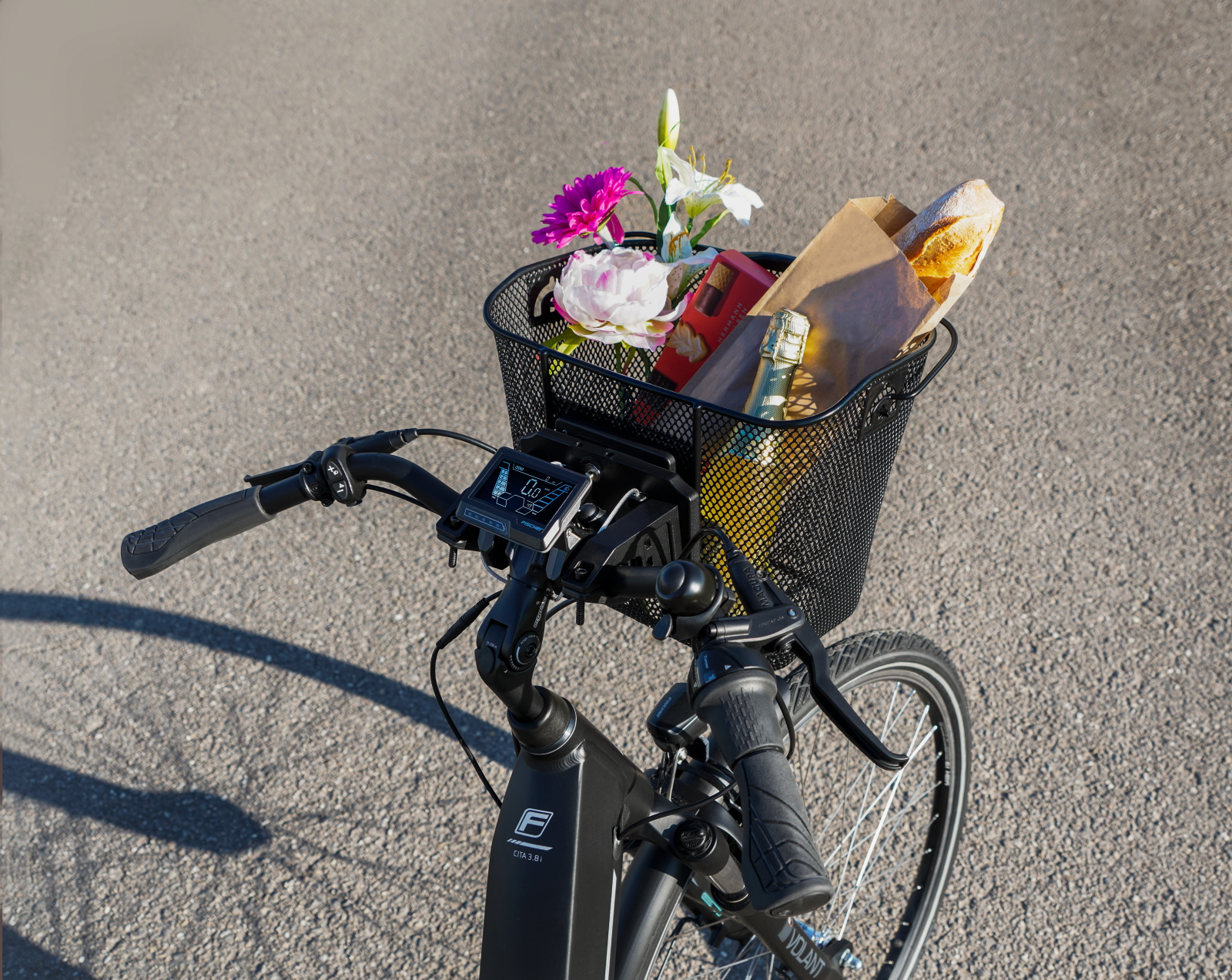 Cesta de bicicleta con flores, baguette y botellas. El manillar de la bicicleta es visible.