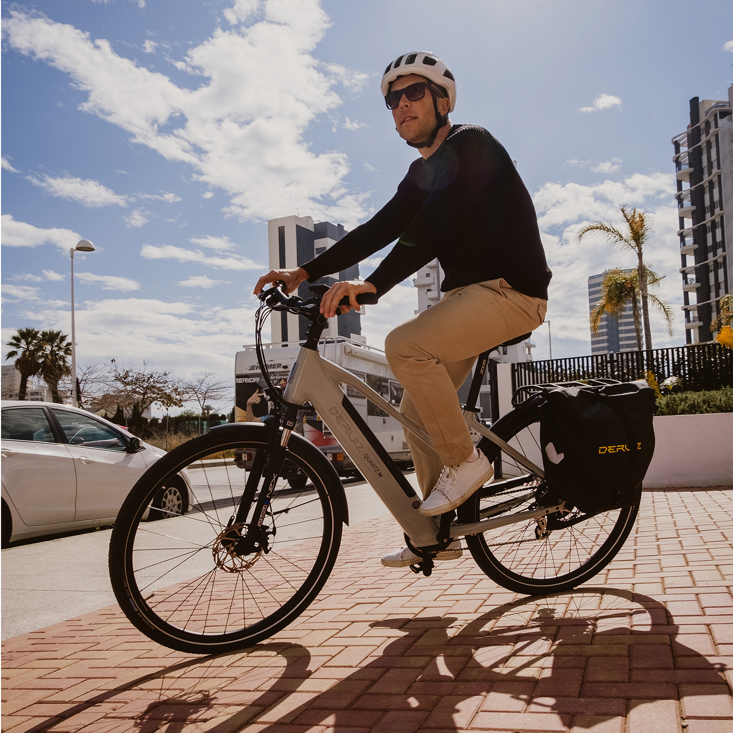 Mann fährt an einem sonnigen Tag in einer Stadt mit einem grauen Deruz E-Bike. Palmen und Gebäude im Hintergrund.