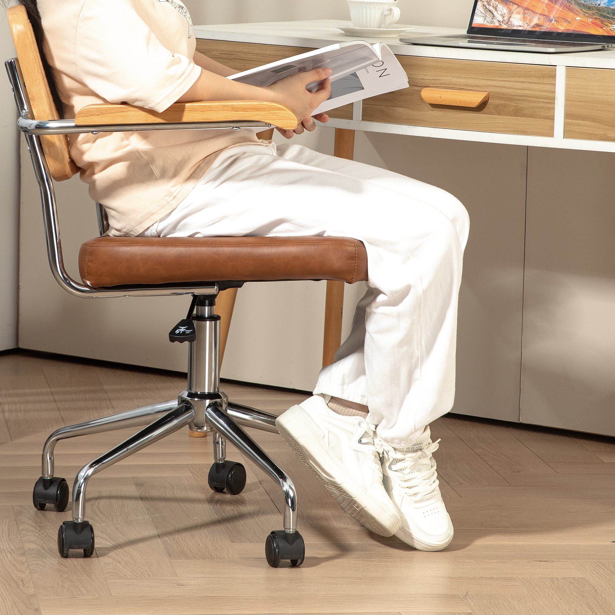 Una persona sentada en un escritorio leyendo un libro. Escritorio y silla de madera, zapatillas blancas.
