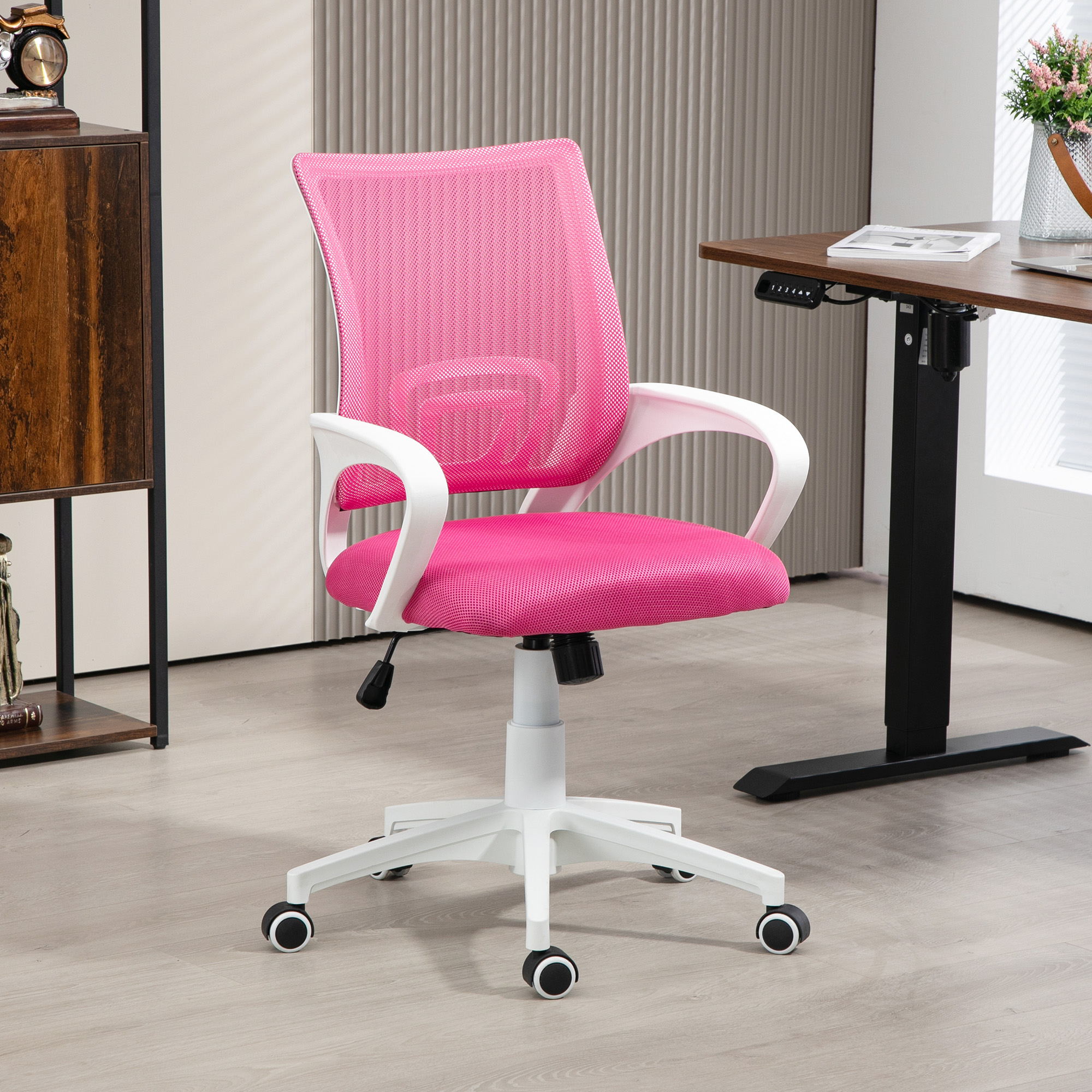 Una silla de oficina rosa con respaldo de malla, brazos y base blancos, cerca de un escritorio marrón y un armario de madera.