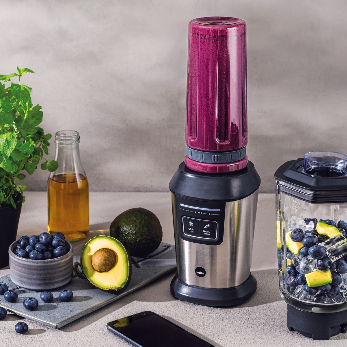 Frullatore con contenuto viola, mirtilli, avocado e telefono su bancone.