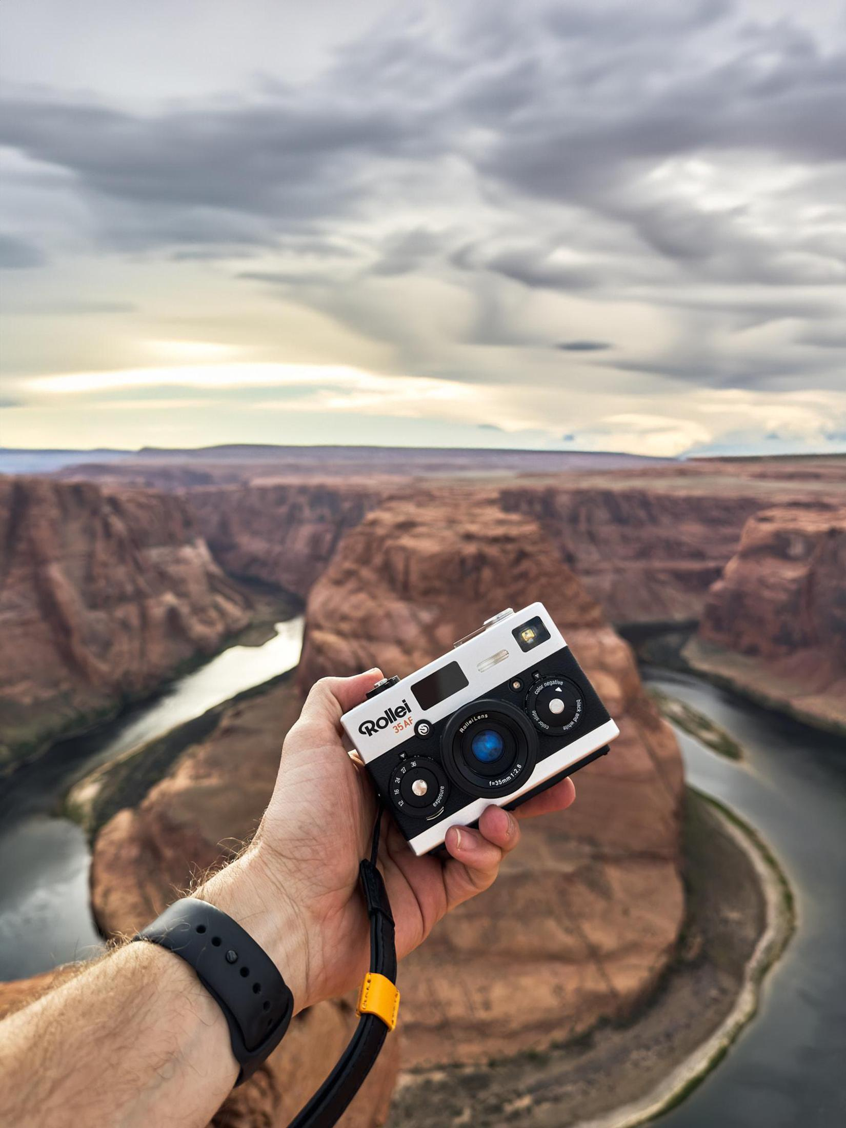Een persoon houdt een zwarte en zilveren Rollei camera vast, tegen een achtergrond van een canyon en bewolkte lucht.