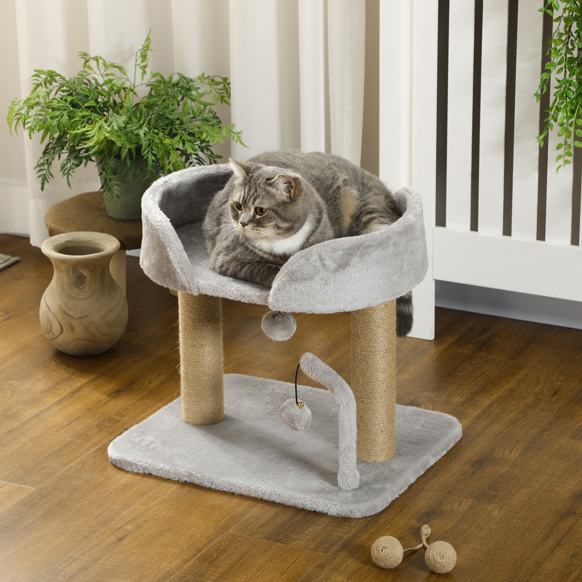 Gatto grigio su un albero per gatti grigio, con un vaso e una pianta verde su pavimento in legno, sfondo bianco.