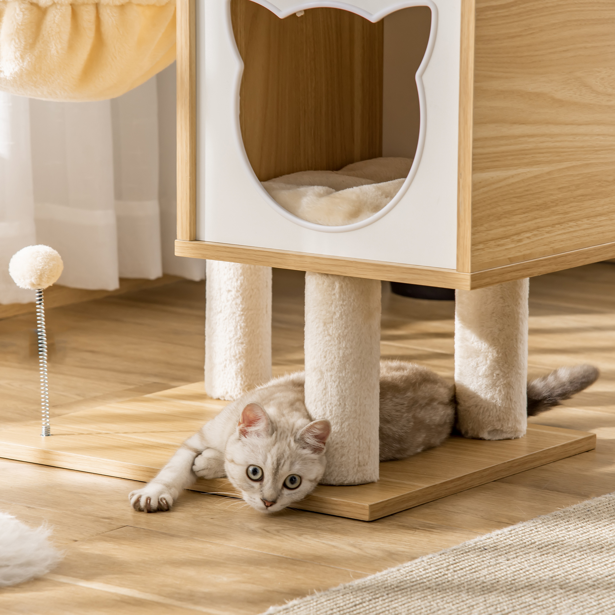 Un gato tumbado bajo un árbol para gatos con casita y un juguete colgante.